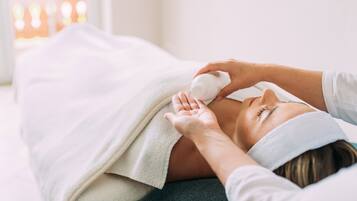 A person lying on a bed with a white blanket, receiving a facial treatment.