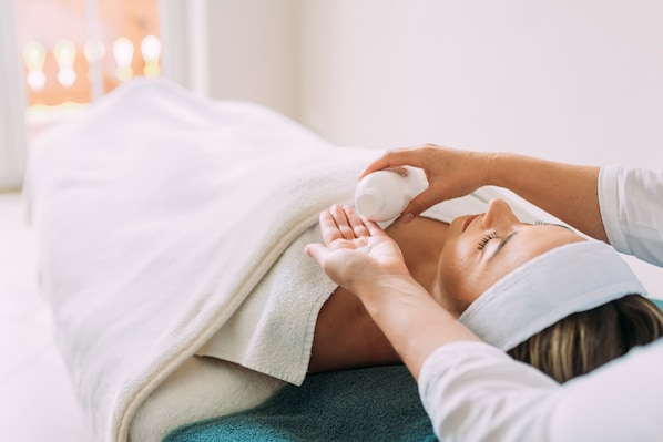 Una persona recostada en una cama con una manta blanca, recibiendo un tratamiento facial.