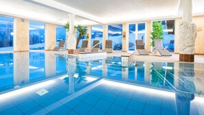 An indoor swimming pool with clear blue water, sun loungers, and large windows offering scenic views. - Naturhotel Reissenlehen (Bischofswiesen)