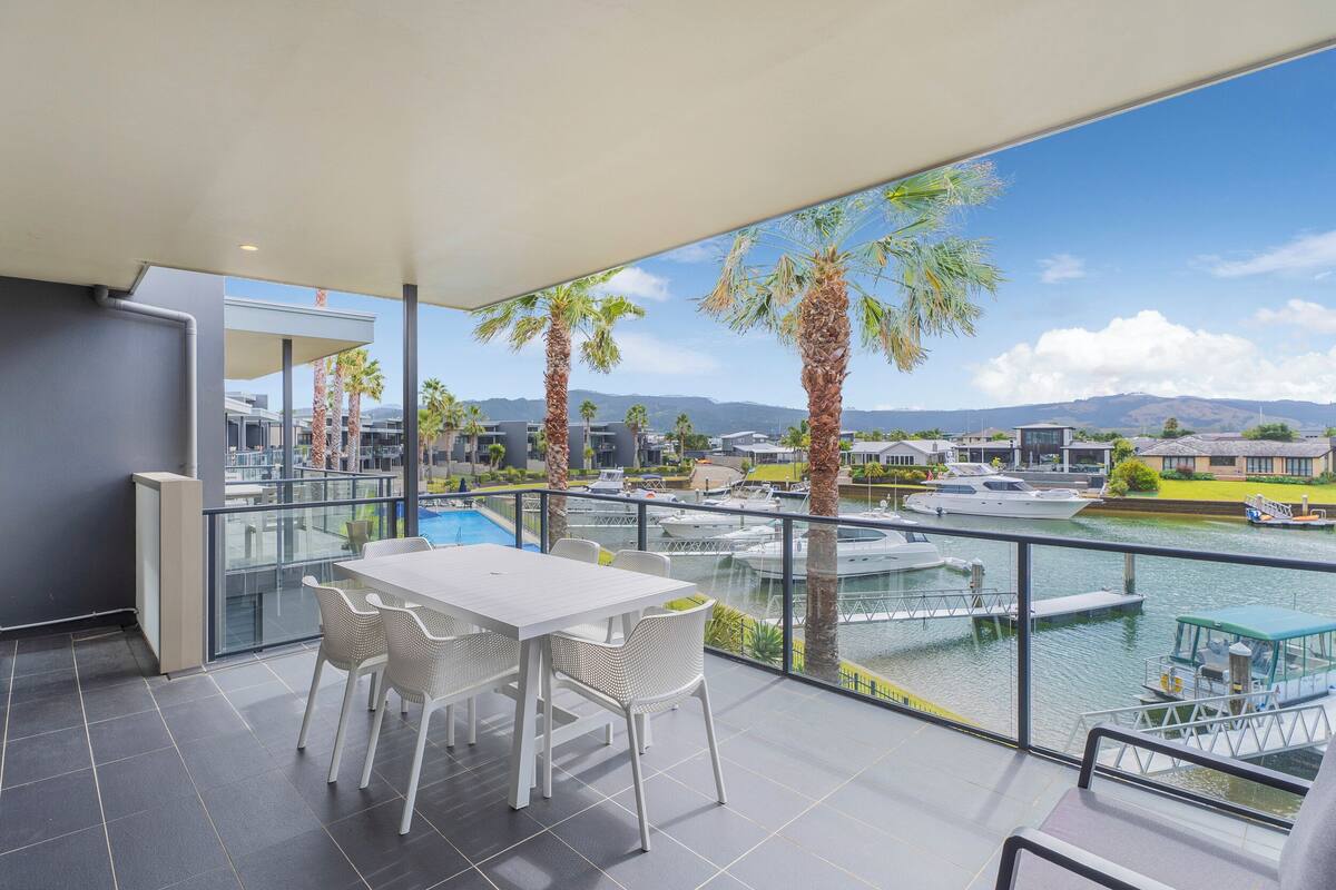 Een balkon met een witte tafel en stoelen, met uitzicht op een jachthaven met boten en palmbomen.