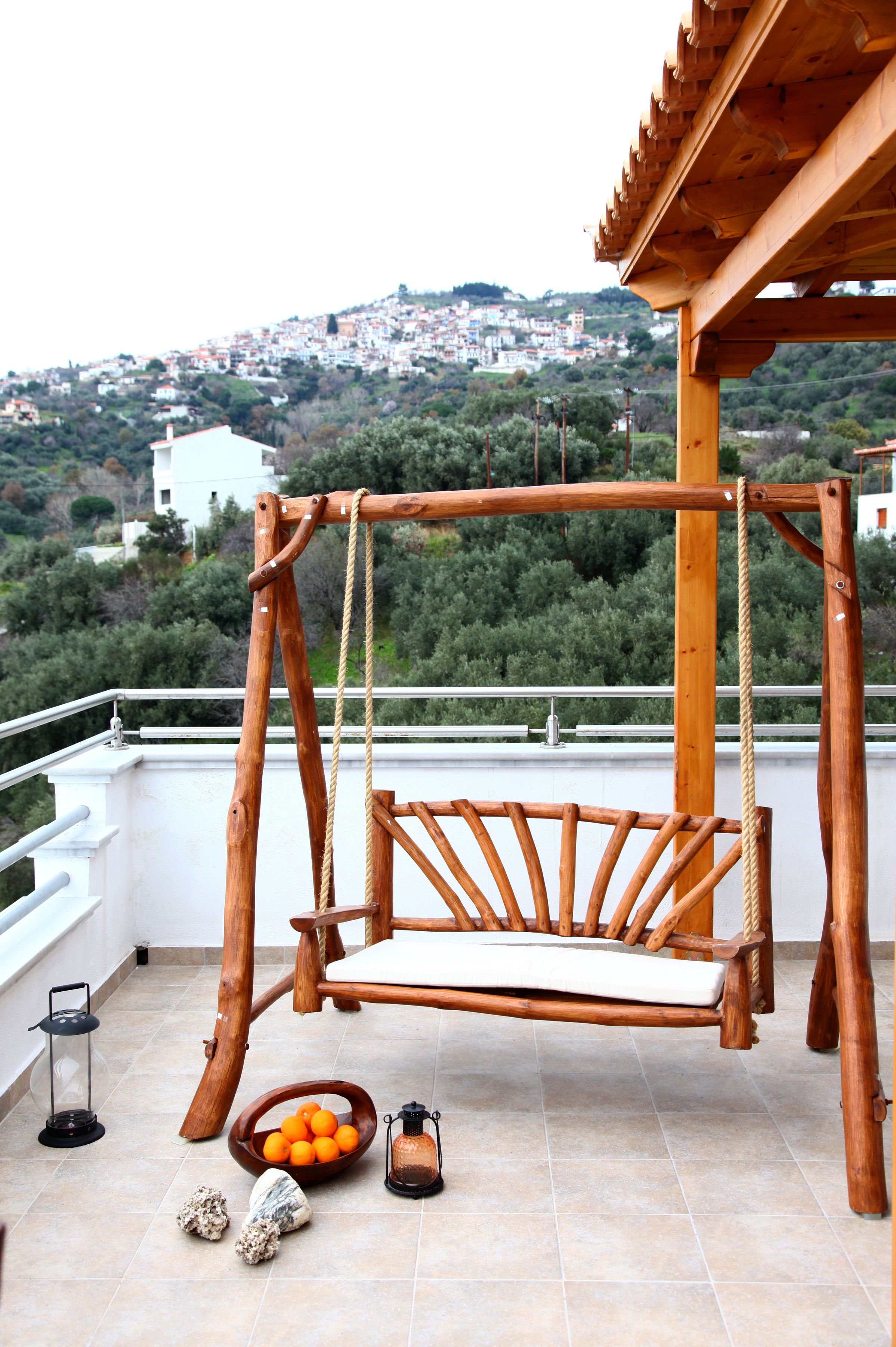 Une chaise suspendue en bois sur un balcon, avec vue sur une ville située sur une colline.