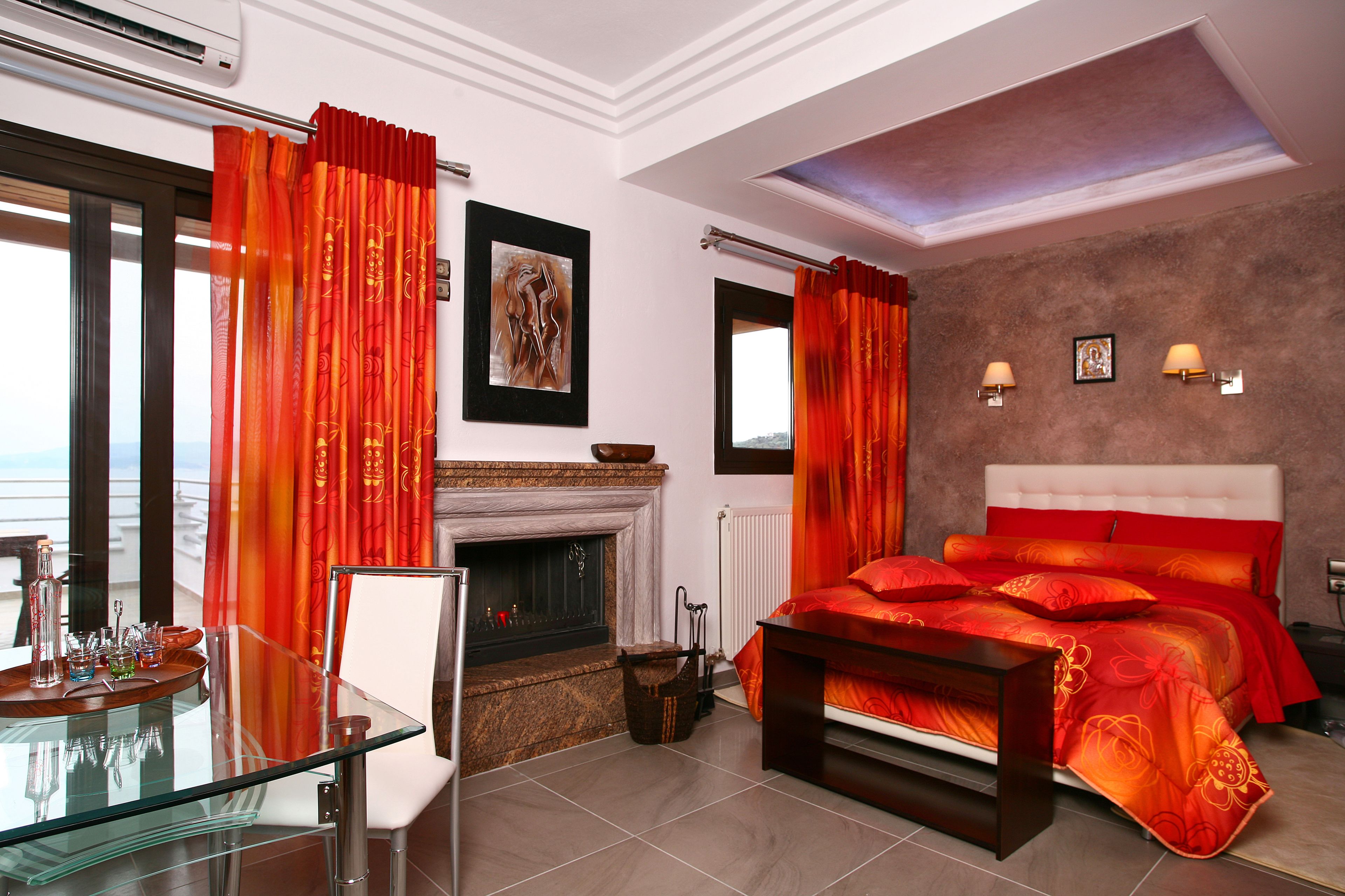 A modern bedroom with a fireplace, a glass dining table, and a bed with red bedding.