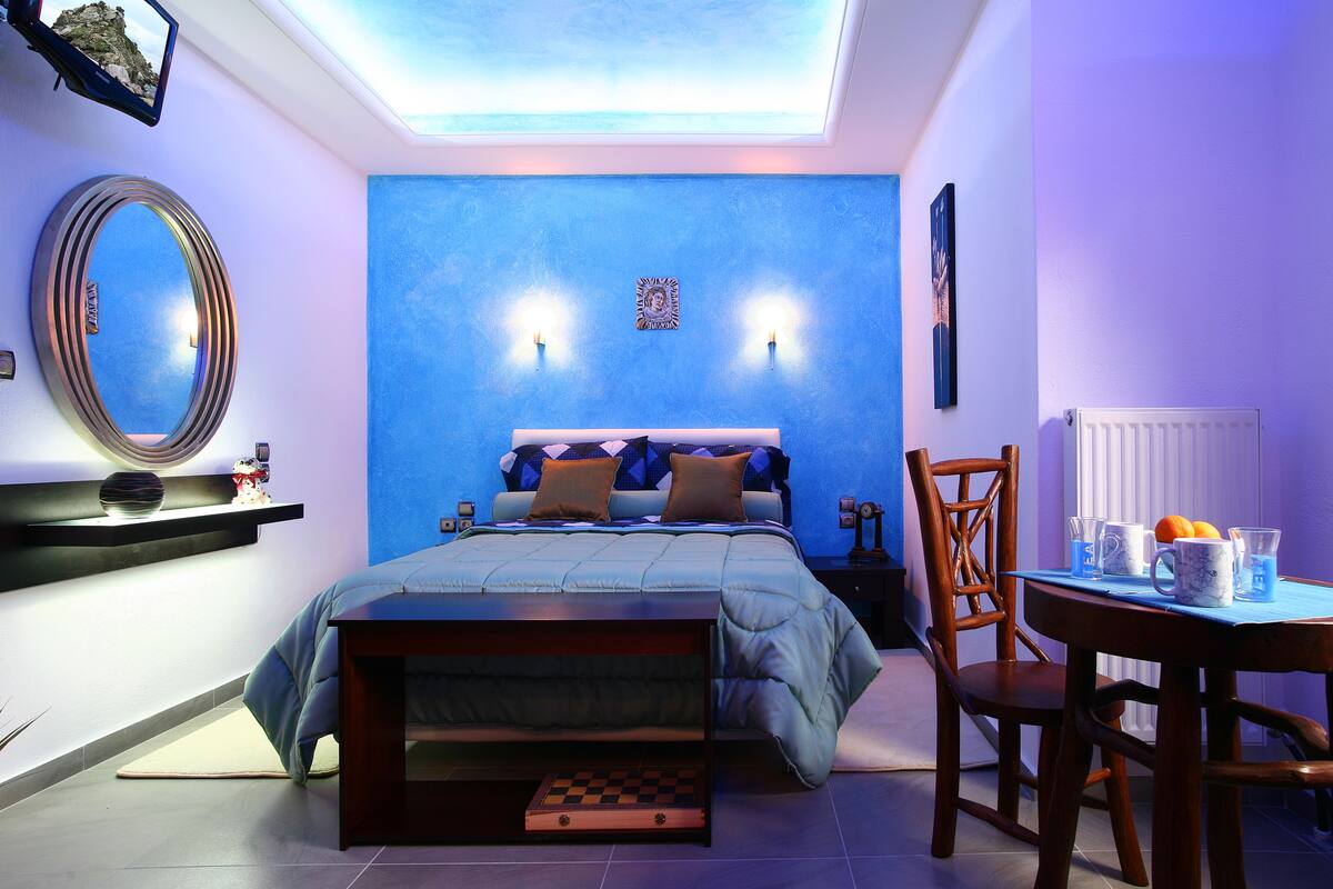 A bedroom with a blue accent wall, a bed with a headboard, bedside tables, a mirror, and a small dining area with a table and chairs.