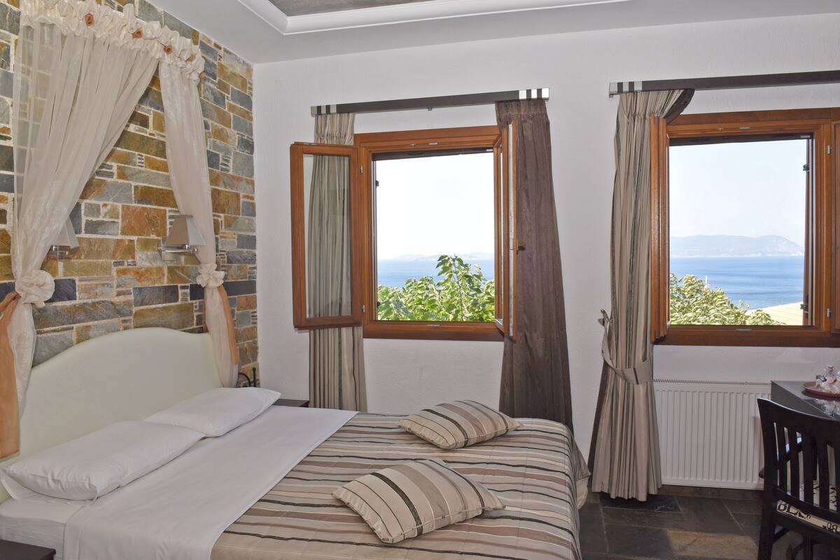 A bedroom with a stone wall, a bed with striped bedding, two windows with curtains, and a view of the sea.