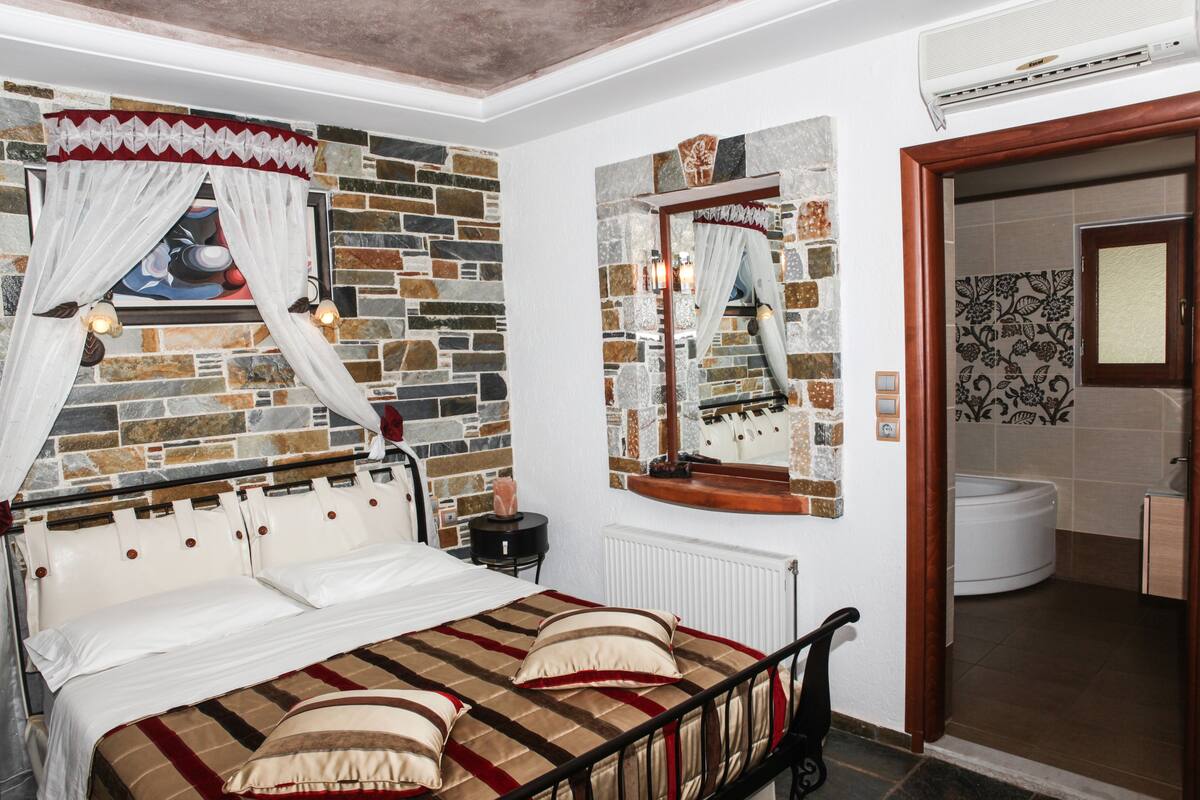 A bedroom with a bed, a stone wall, a mirror, and a bathroom visible through an open door.