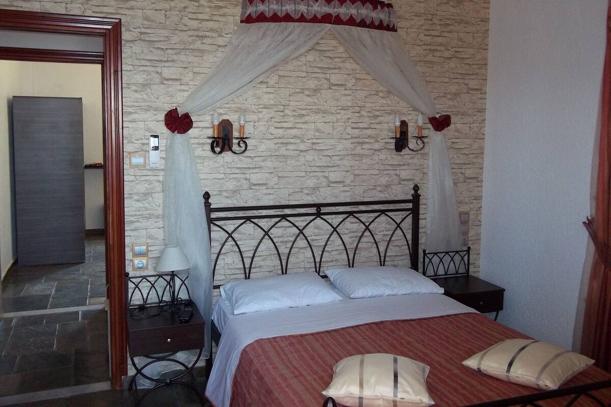 A bedroom with a stone wall, a bed with white linens, a red-striped bedspread, a nightstand with a lamp, and a decorative canopy.