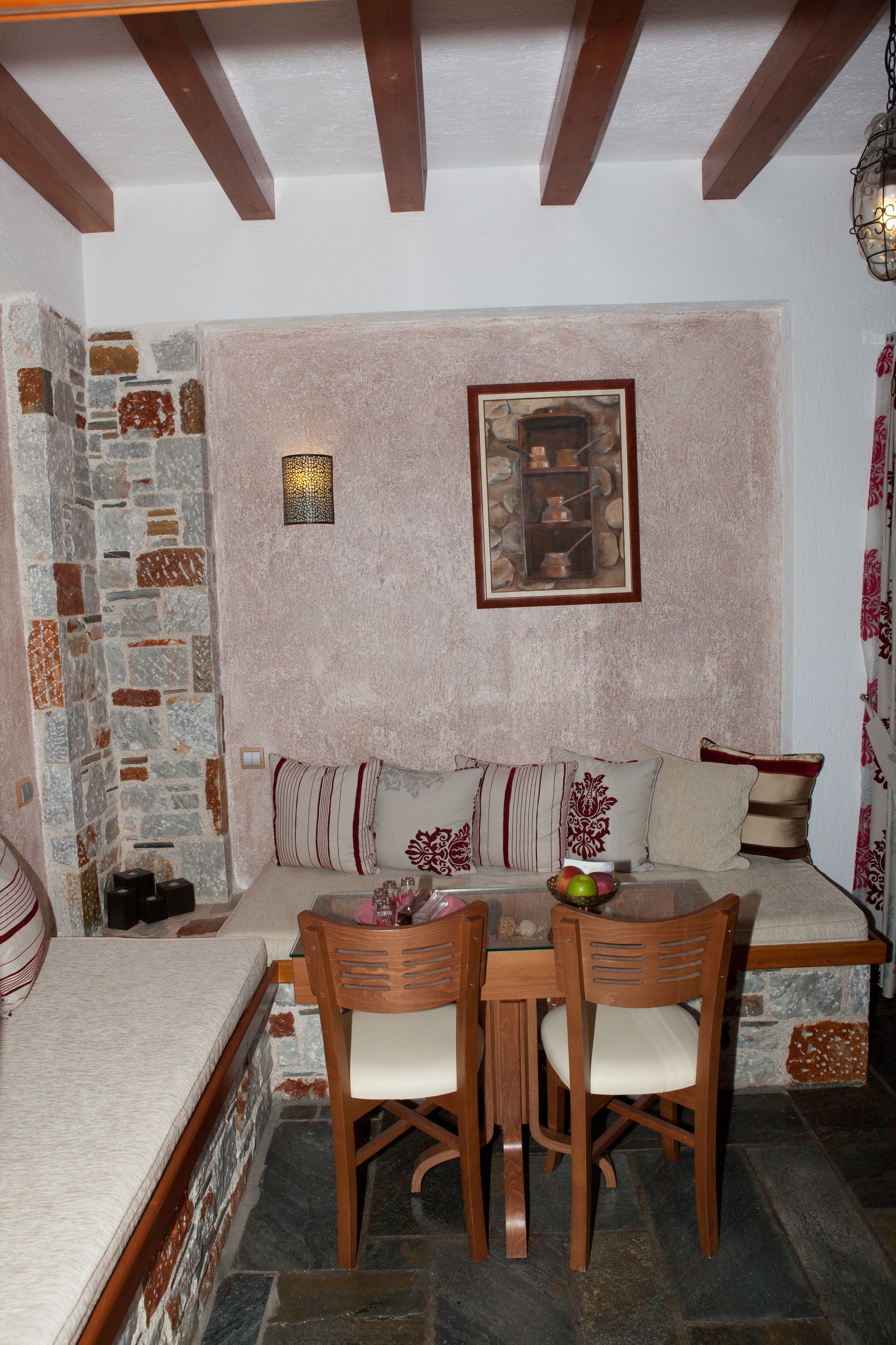 A cozy room with a stone wall, wooden ceiling beams, a cushioned bench, a table with chairs, and a framed picture on the wall.
