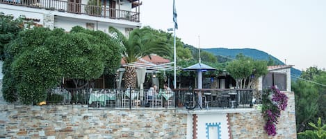 Un edificio de piedra con balcón, mobiliario exterior y montañas al fondo.