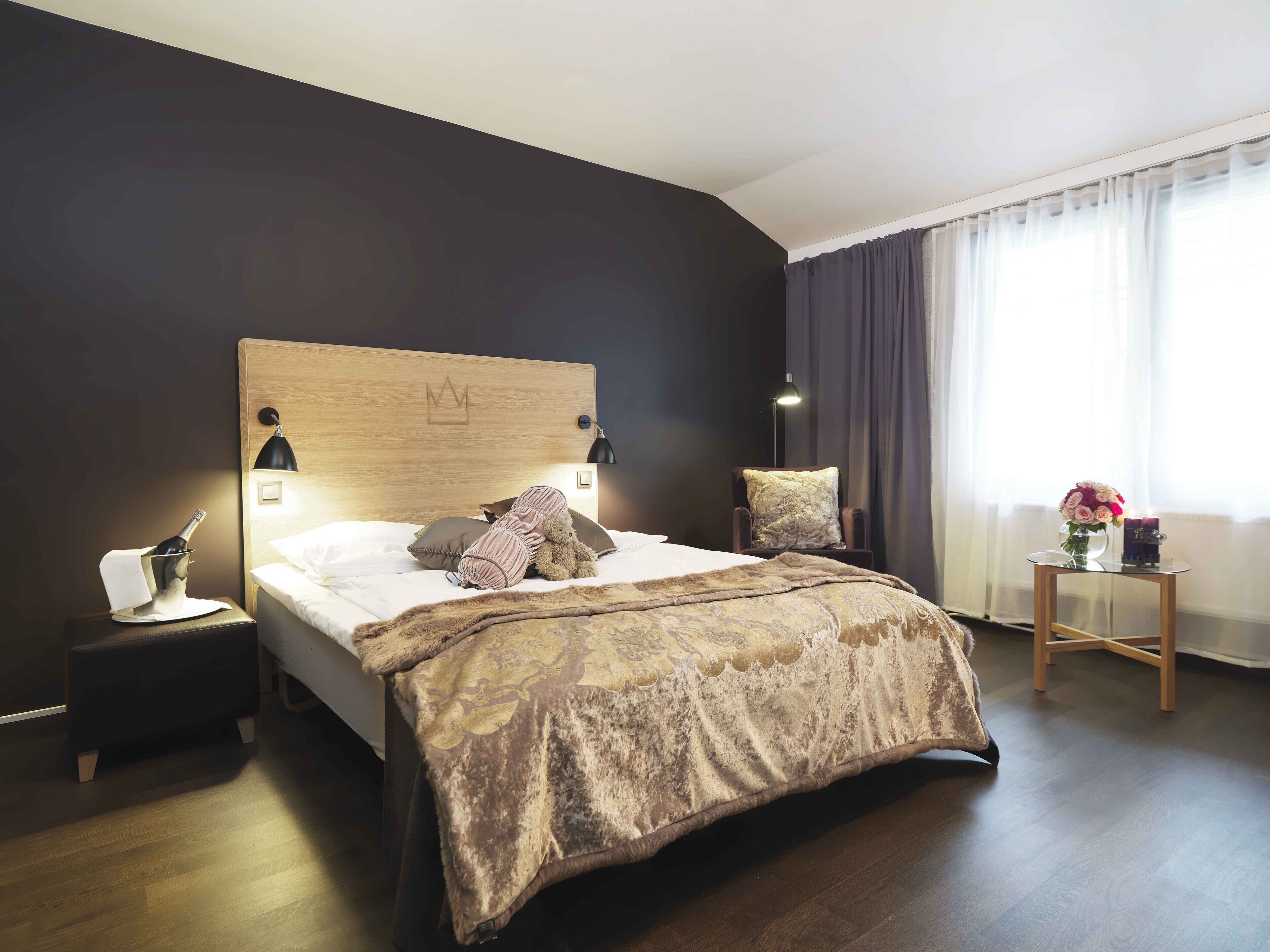 Hotellihuone, jossa on suuri sänky, yöpöytä, jossa on samppanjapullo, pehmustettu tuoli, pieni pöytä, jossa on maljakko täynnä kukkia, ja suuri ikkuna, jossa on verhot.