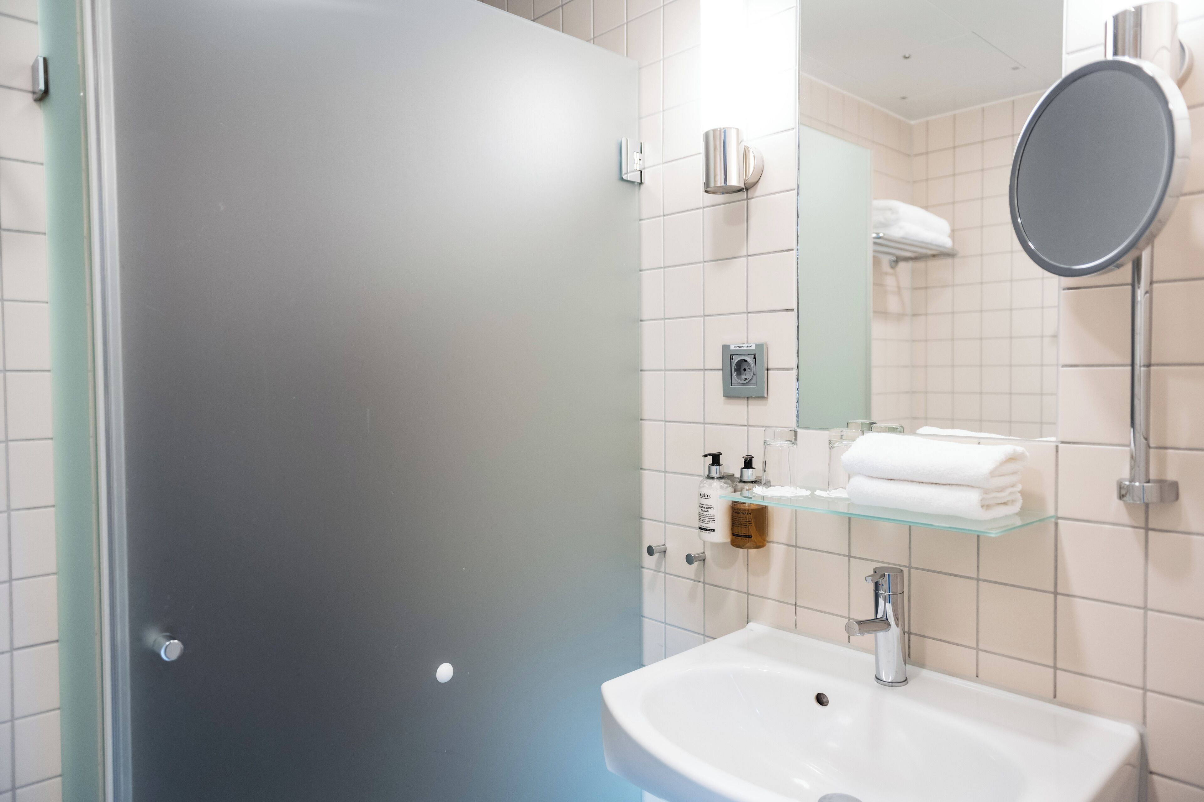 Et moderne badeværelse med brusekabine i glas, et hvidt håndvaskområde med spejl, og pænt arrangeret toiletartikler på en hylde.