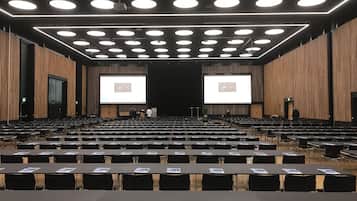 A large conference room with rows of tables and chairs, a stage with a large screen, and a modern ceiling design.
