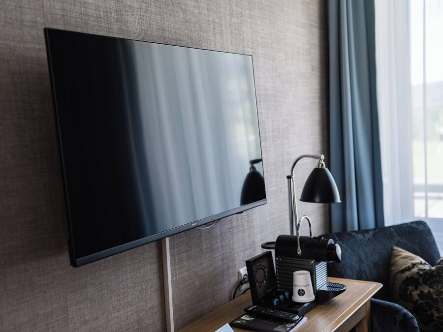 A modern hotel room with a large flat-screen TV mounted on the wall, a desk with a lamp, a coffee maker, and a remote control.