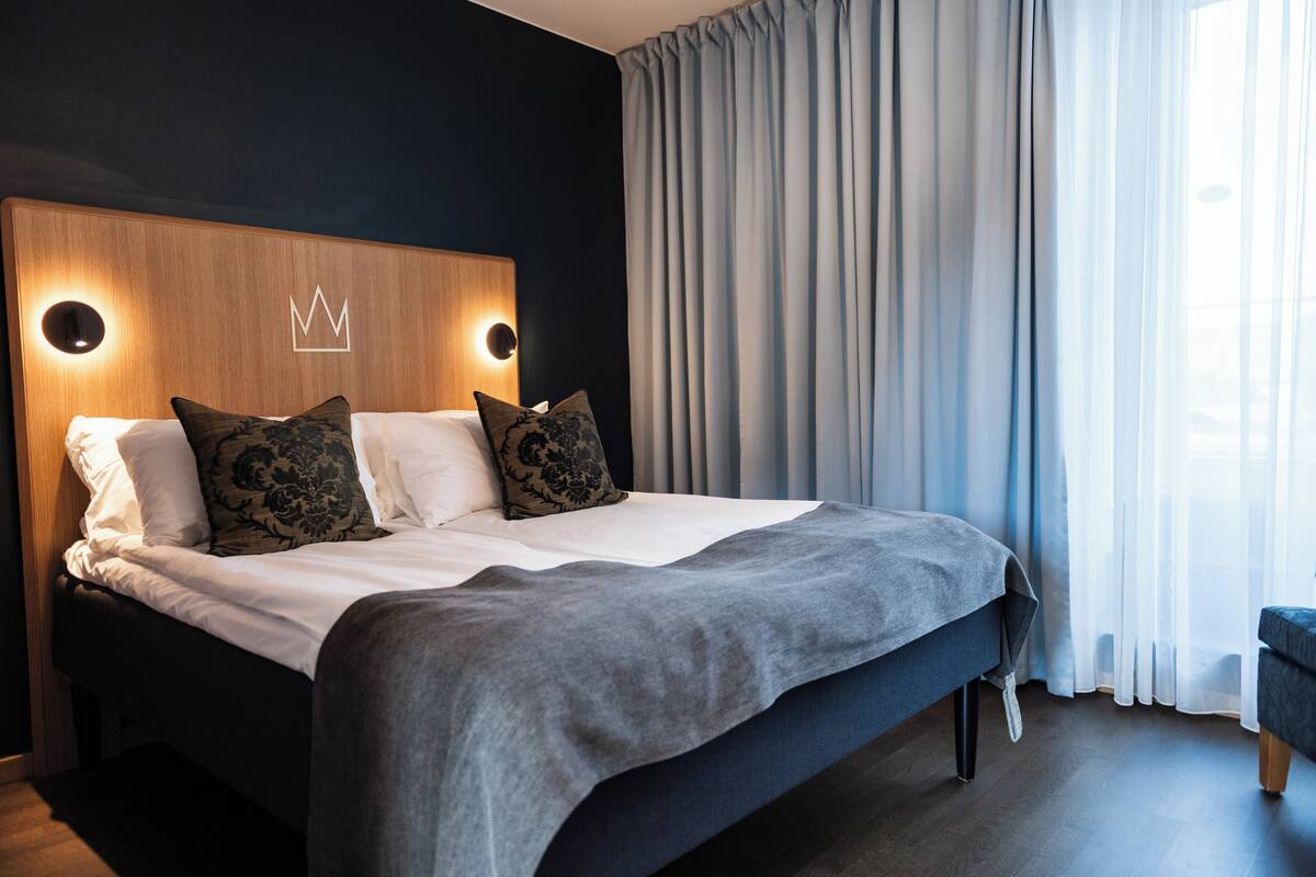 Una habitación de hotel moderna con una cama grande, cabecera de madera con iluminación en forma de corona y cortinas translúcidas.