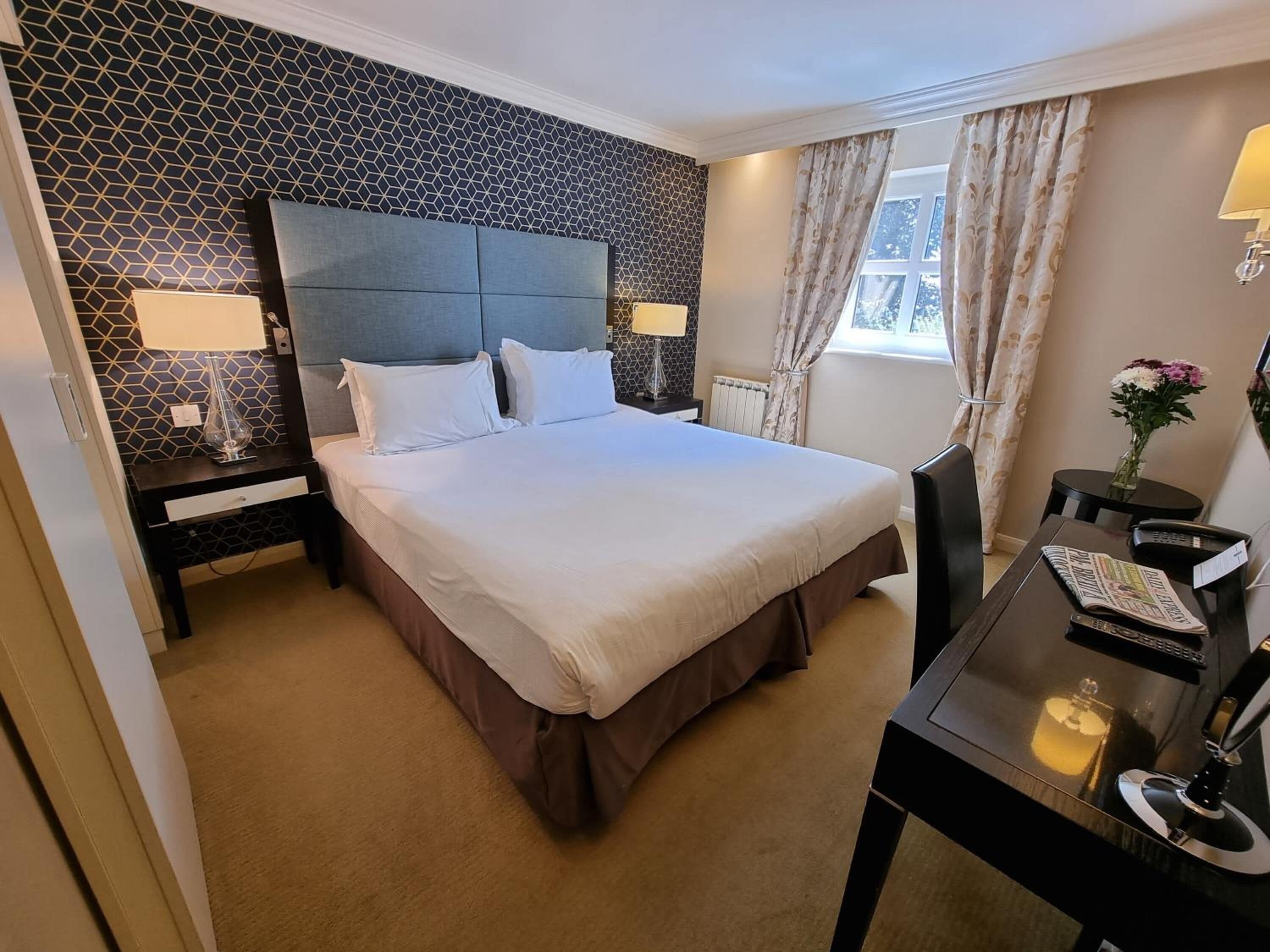 A hotel room with a large bed, two bedside tables with lamps, a desk with a chair, and a window with curtains.