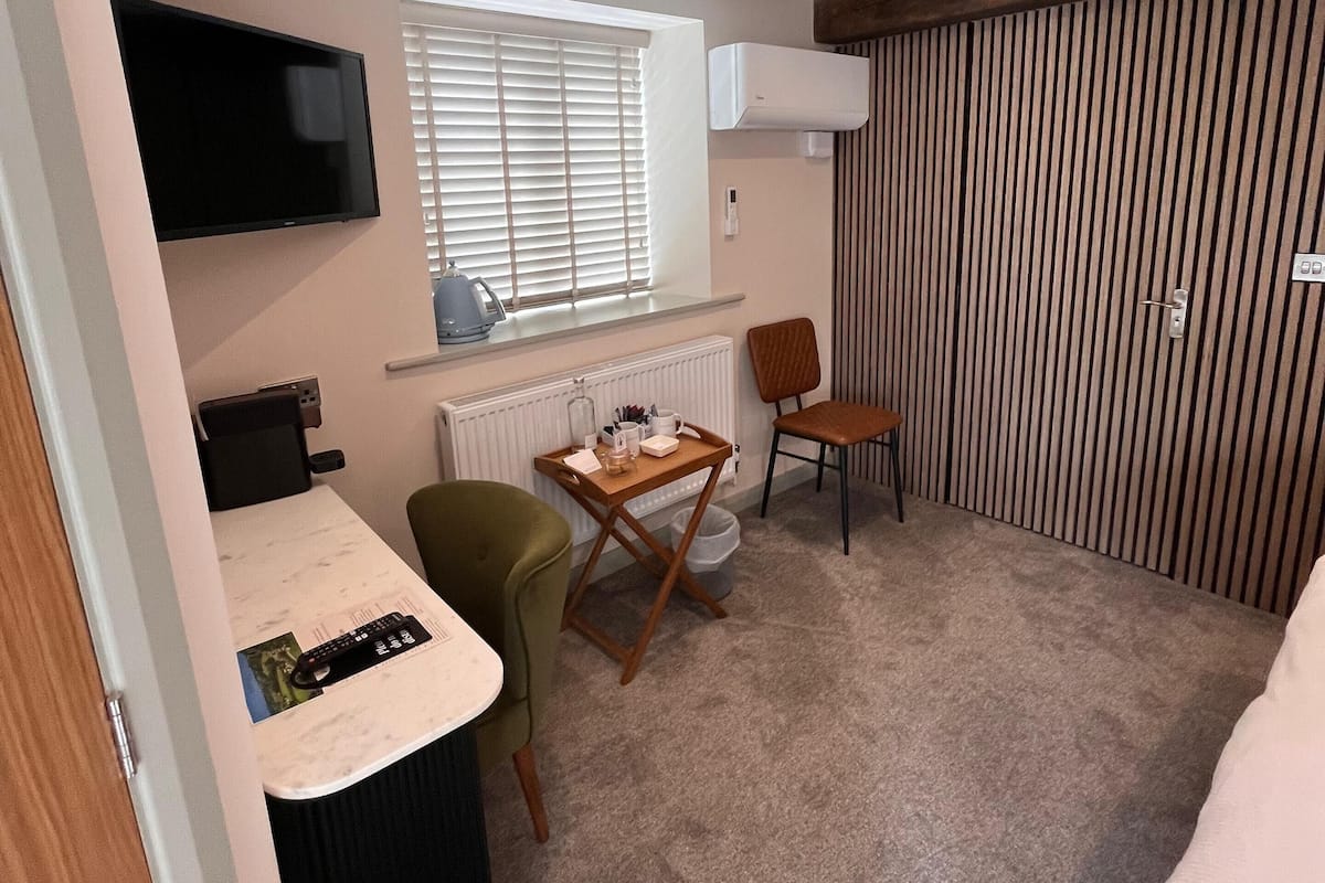 Quarto compacto com uma cama, escrivaninha com cadeira, televisão e janela com persianas.