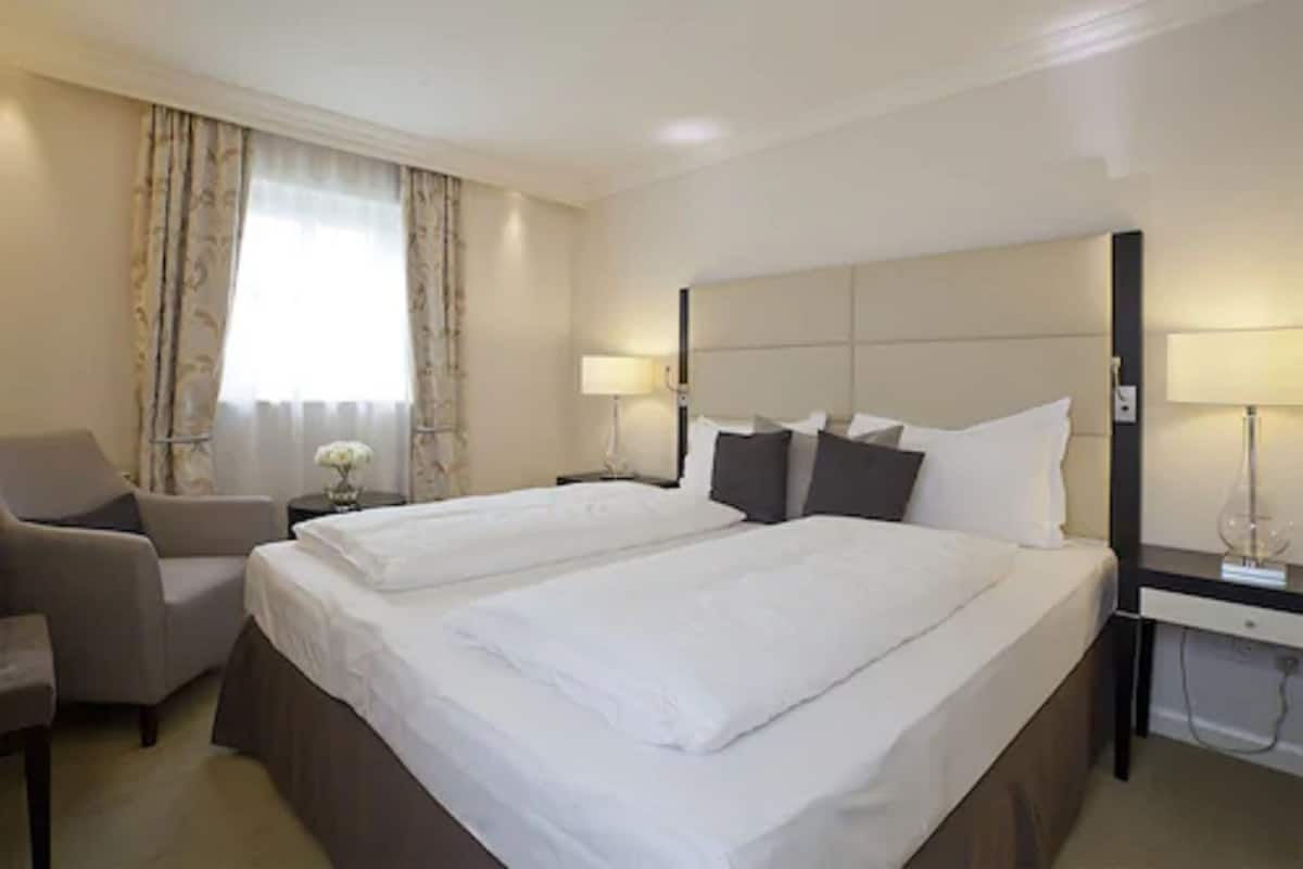 Quarto de hotel com duas camas, uma escrivaninha, uma cadeira e uma janela com cortinas.