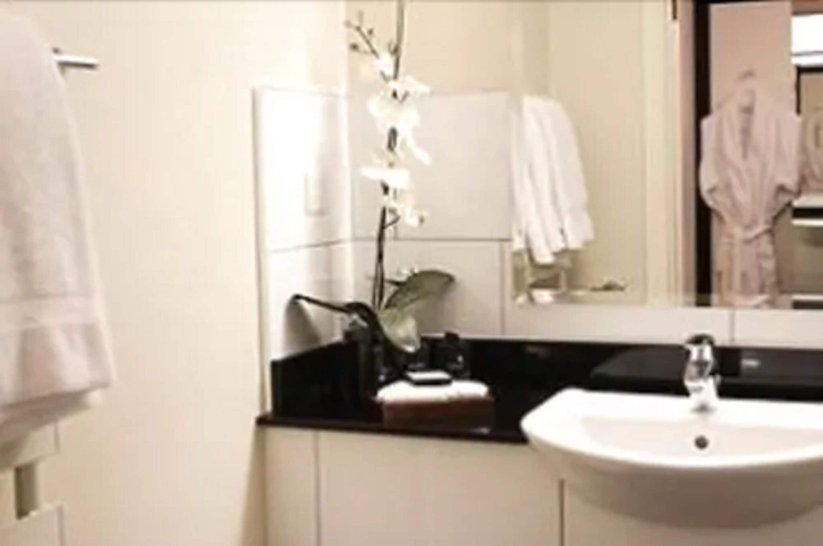 Une salle de bain avec un lavabo blanc, un plan de toilette noir, une orchidée blanche et un miroir qui reflète une robe blanche.