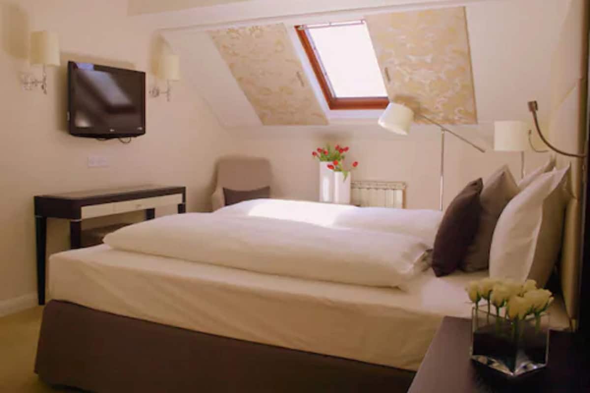 Quarto com cama, escrivaninha, cadeira, televisão, janela e um vaso com flores.