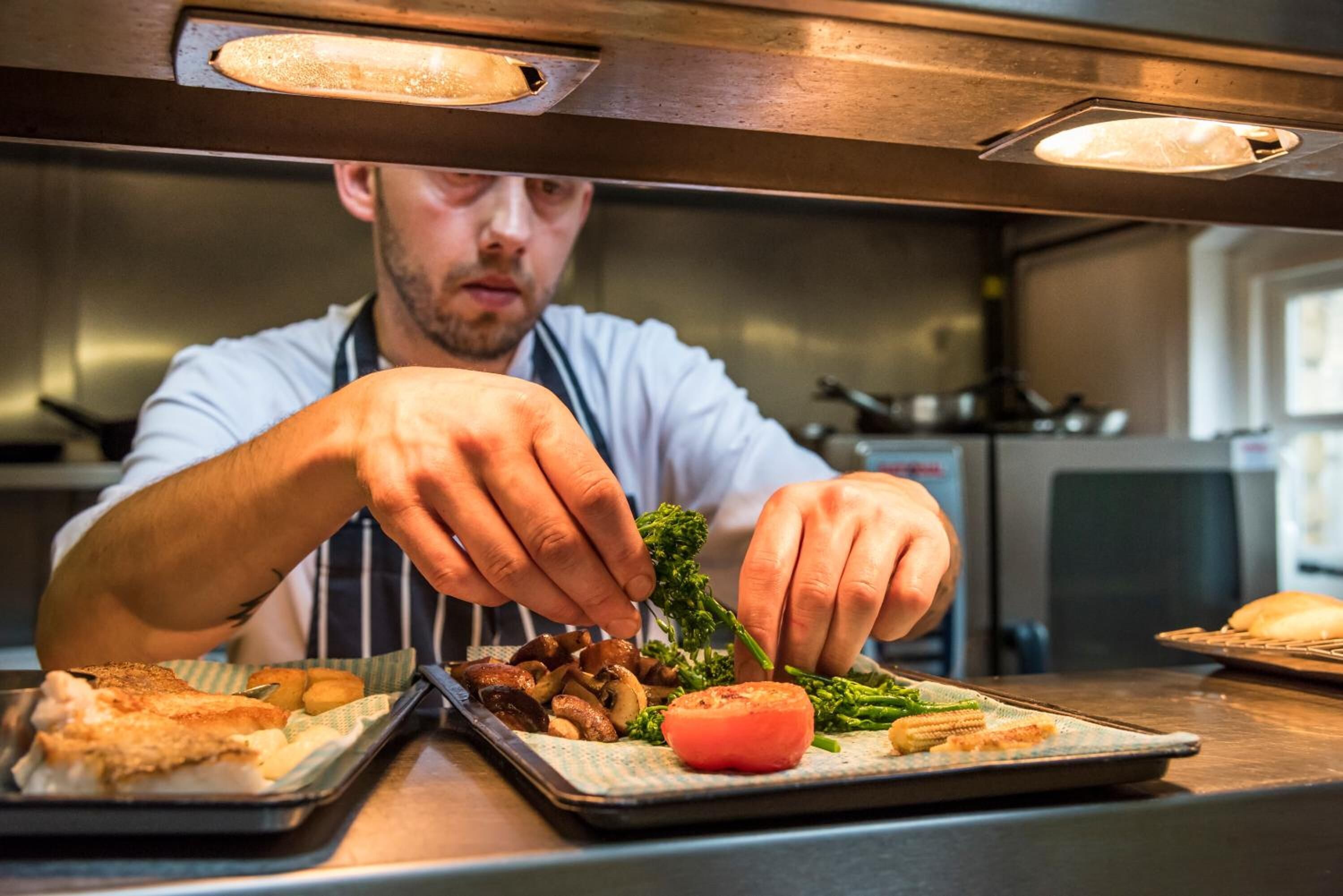 Un chef prépare des plats dans une cuisine équipée d’appareils en acier inoxydable, et son attention se porte sur un plateau contenant des aliments, notamment des tomates, des champignons et des légumes verts.