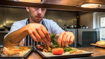Un chef prépare des plats dans une cuisine équipée d’appareils en acier inoxydable, et son attention se porte sur un plateau contenant des aliments, notamment des tomates, des champignons et des légumes verts.