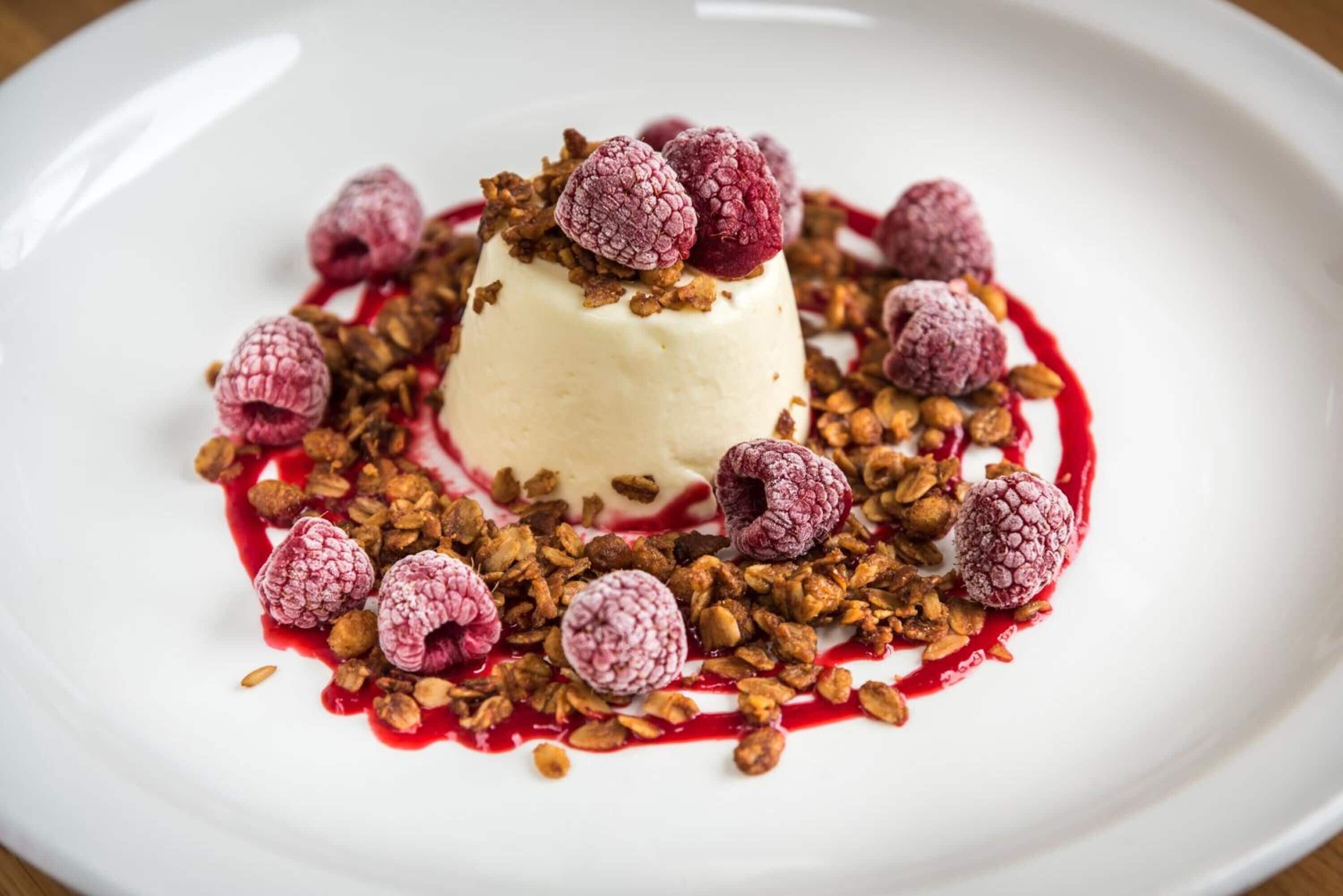 Un dessert servi dans une assiette blanche, garni de framboises et de granola.