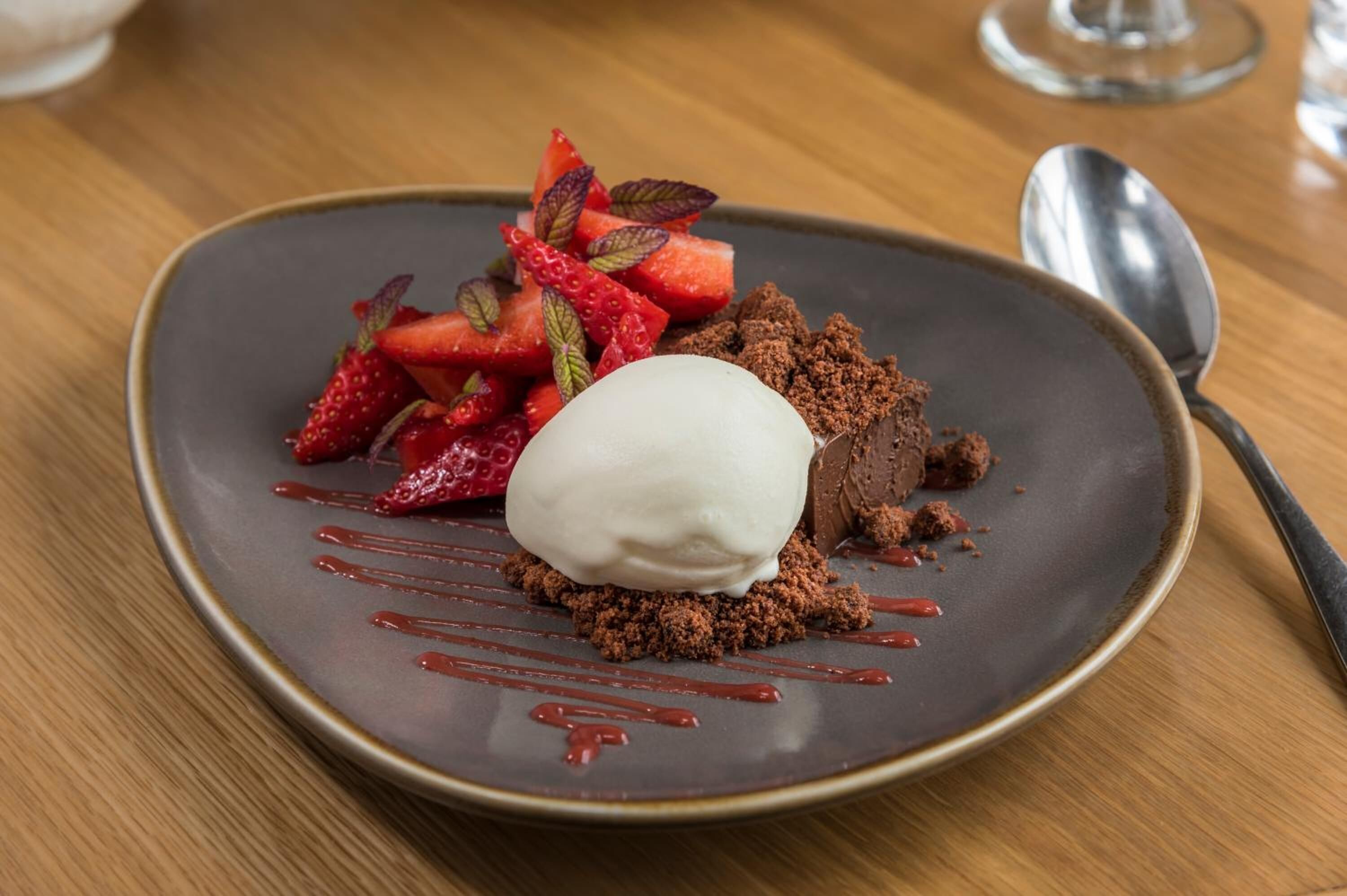 A dessert plate with chocolate crumble, strawberries, and a scoop of ice cream on a dark plate.