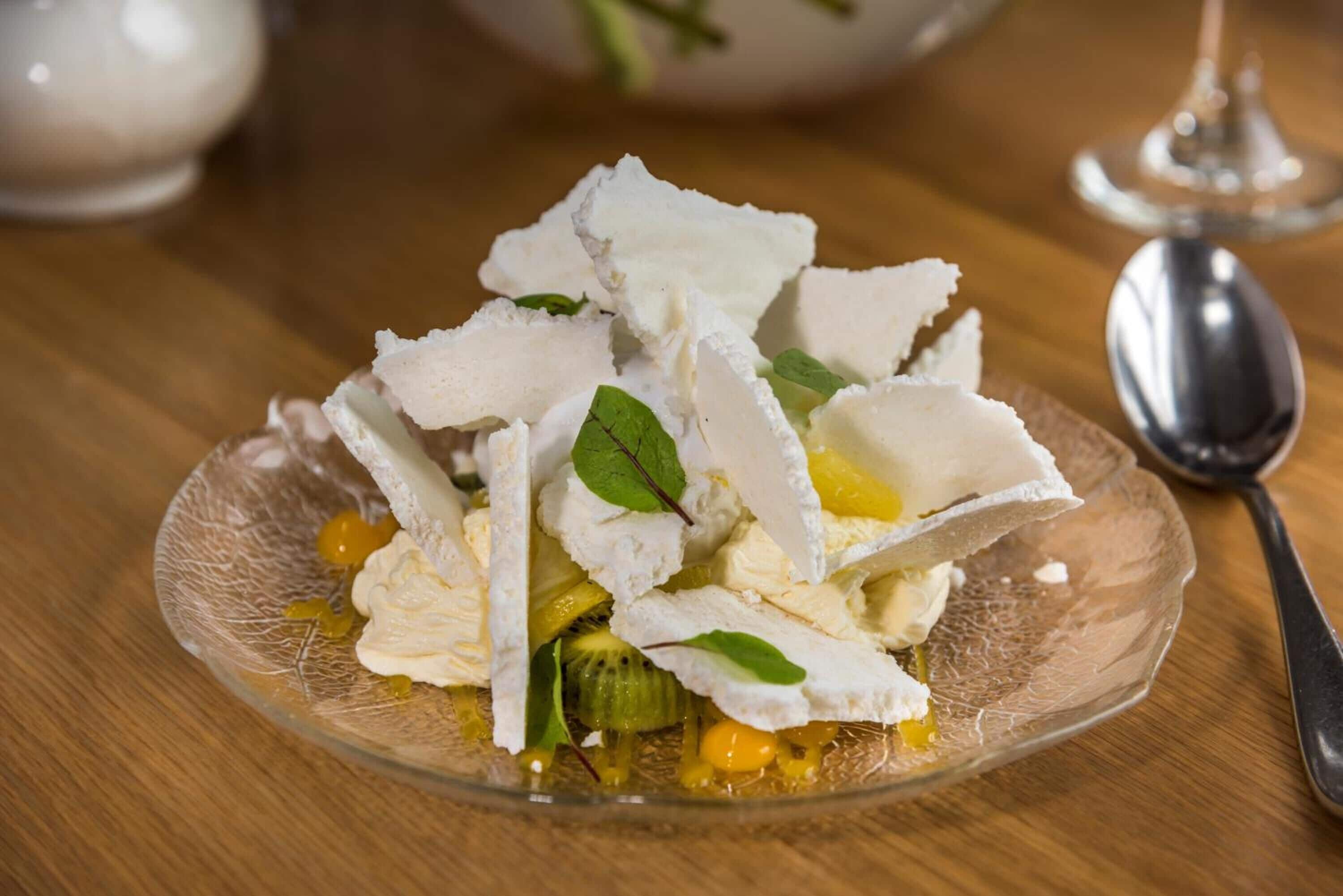 A clear glass bowl containing a salad with white cheese, green leaves, and yellow fruit pieces on a wooden table.