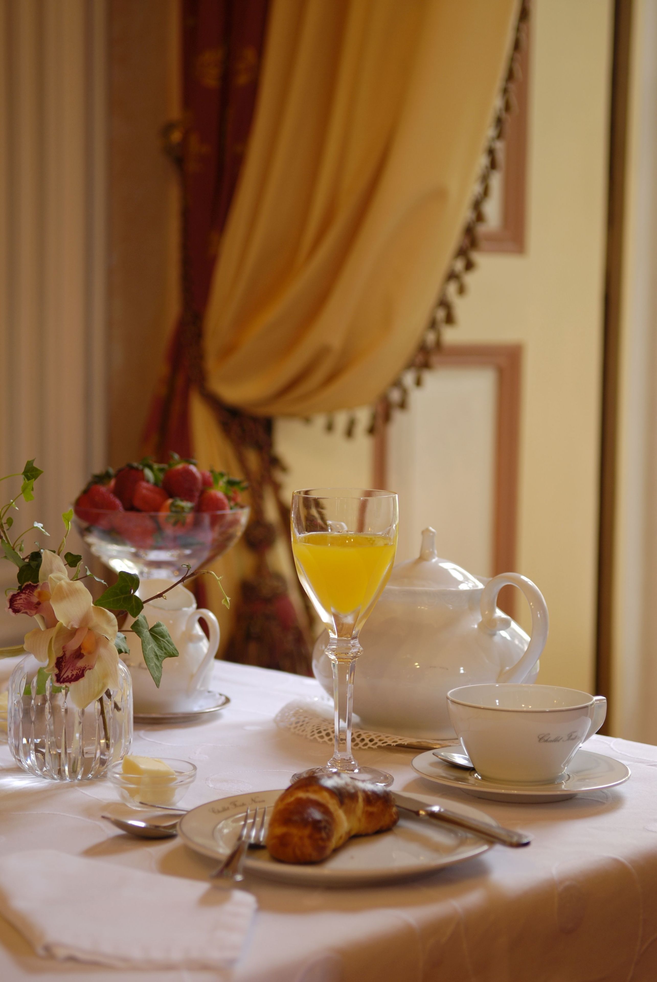 A breakfast table with a teapot, a glass of orange juice, a bowl of strawberries, a plate with a croissant, and a vase with flowers.
