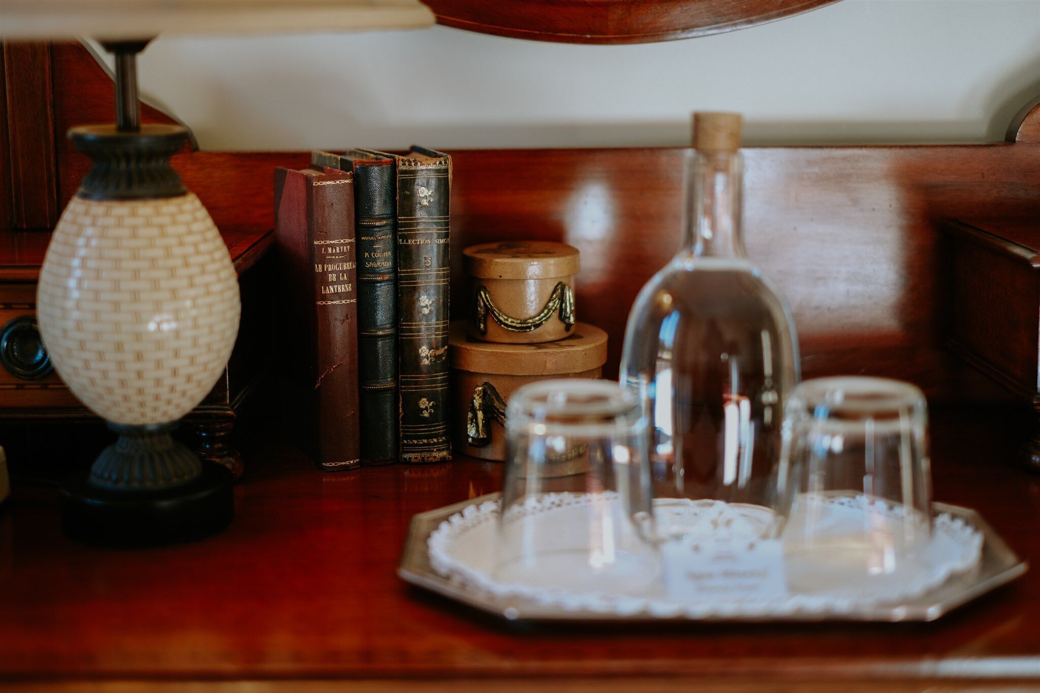 A wooden table with a lamp, books, a glass decanter, and a tray with glasses.