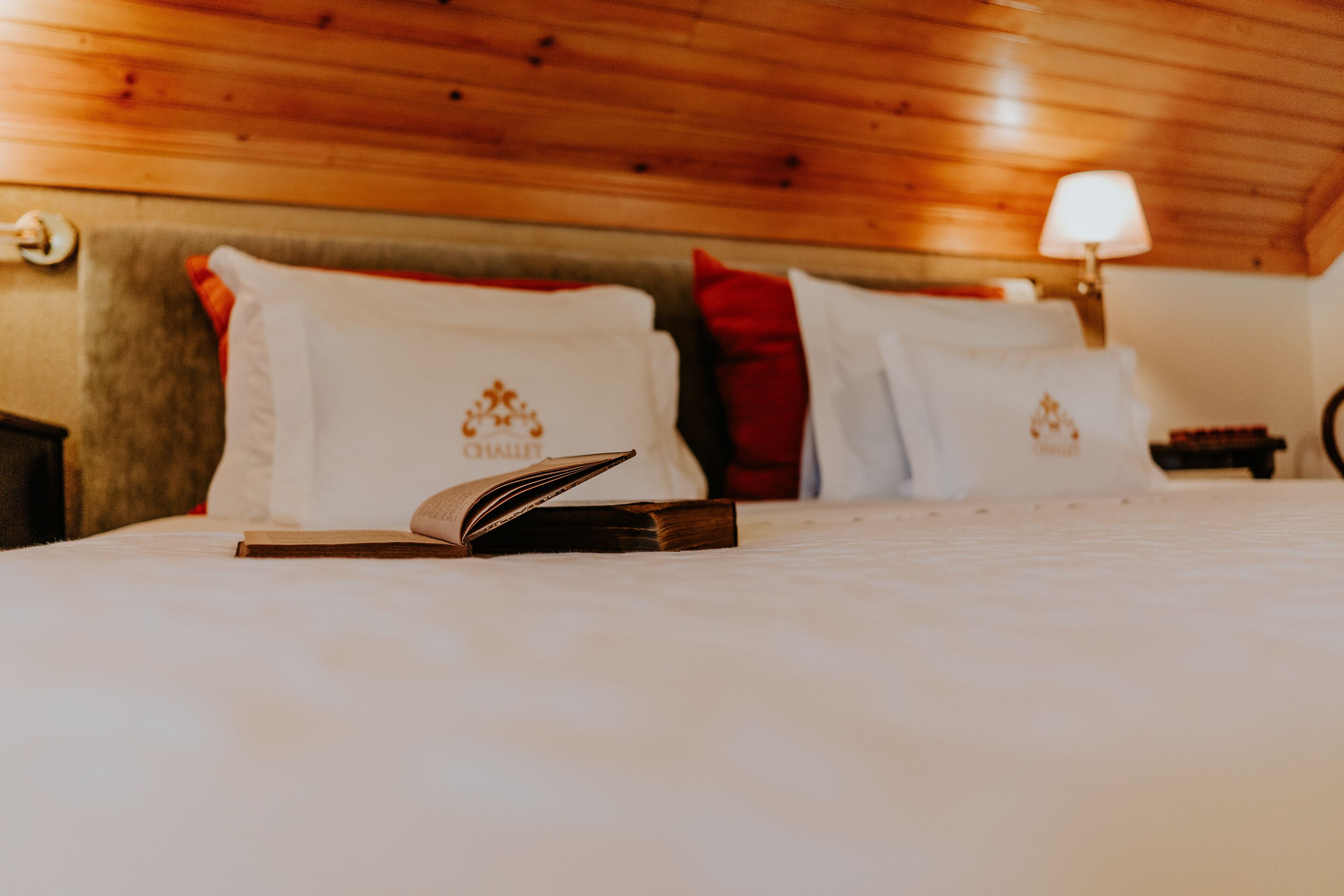Una cama bien hecha con un libro, almohadas con un logotipo y una lámpara en un techo de madera.