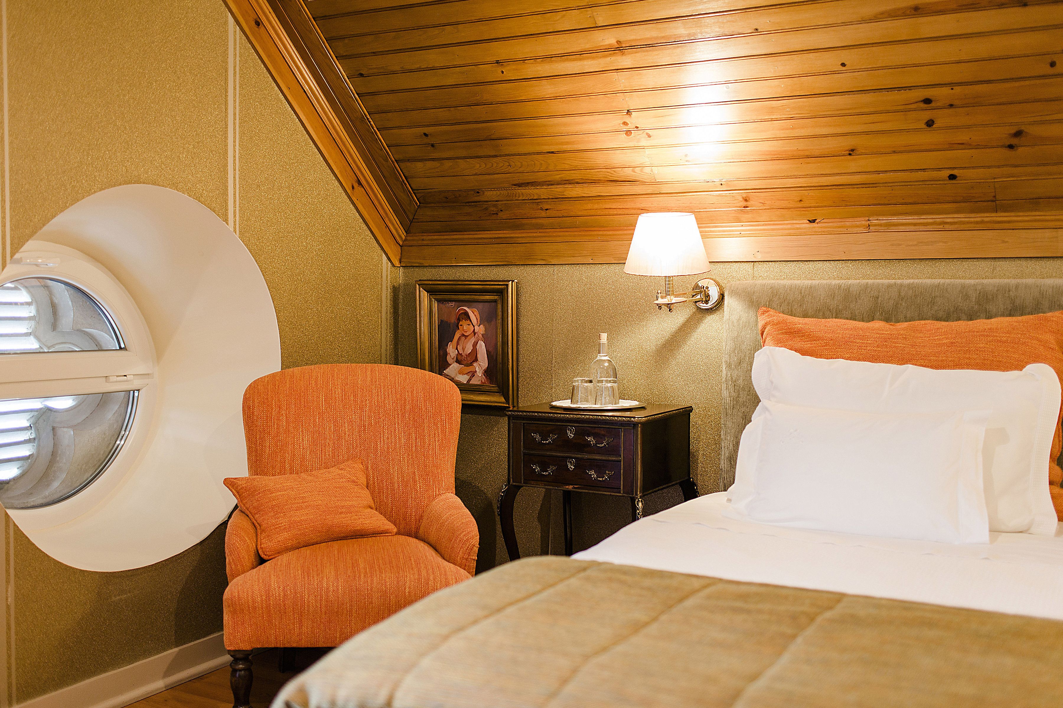 A hotel room with a bed, an orange armchair, a nightstand with a lamp, a picture on the wall, and a window with blinds.