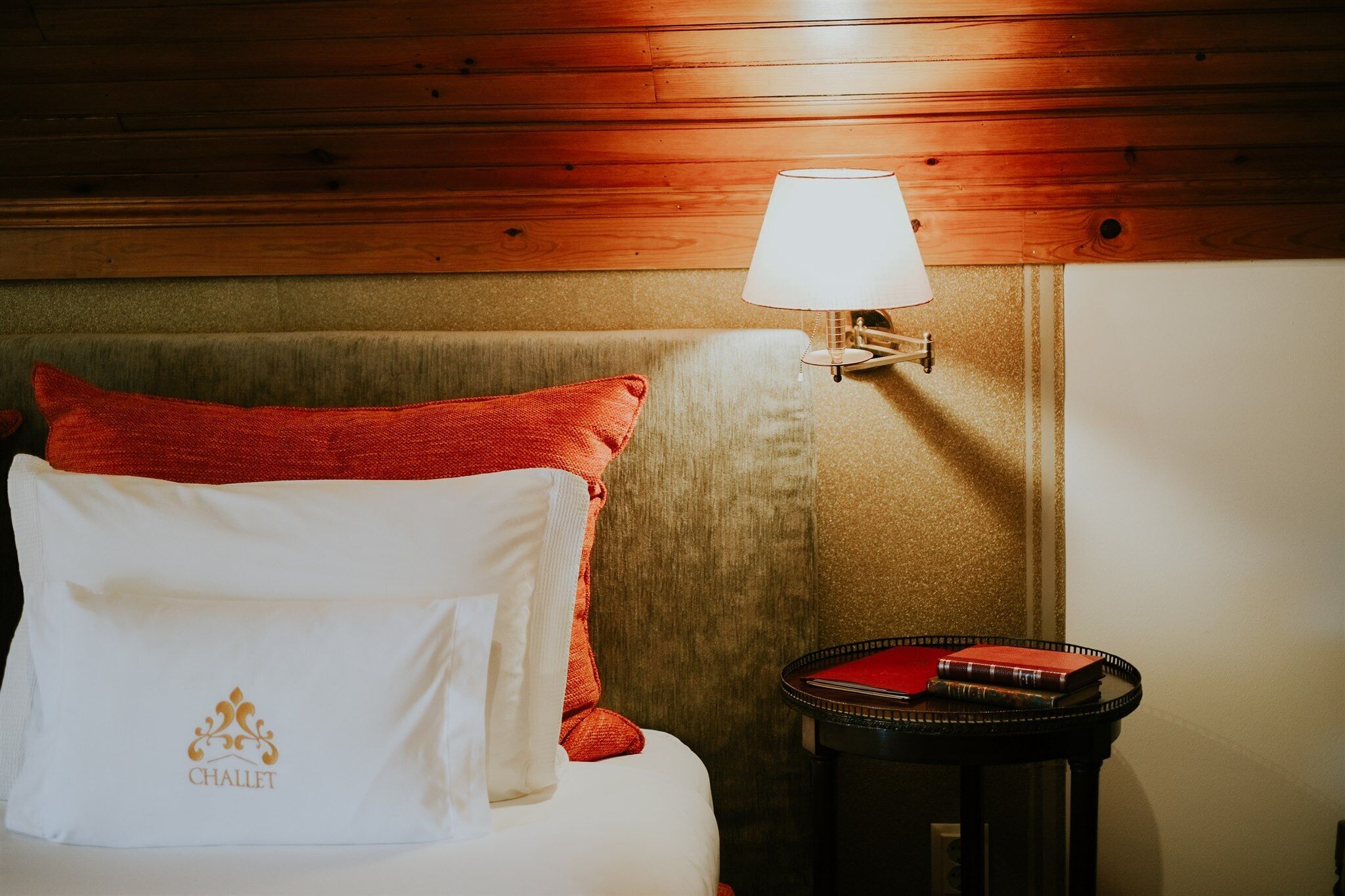 Una habitación de hotel con cabecera de madera, cama con almohadas rojas y blancas, una mesa con lámpara y un pequeño libro rojo sobre la mesa.
