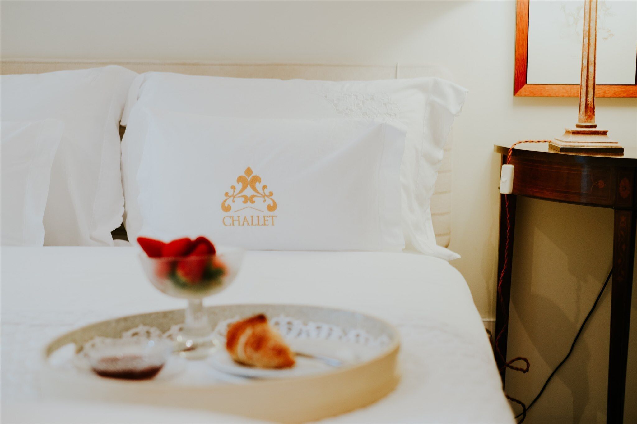 Una cama bien hecha con sábanas blancas y una almohada con un logotipo. En la cama hay un plato con un postre y una copa.