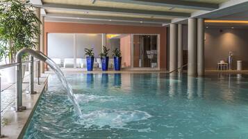 An indoor swimming pool with a clear water feature, surrounded by columns and potted plants.