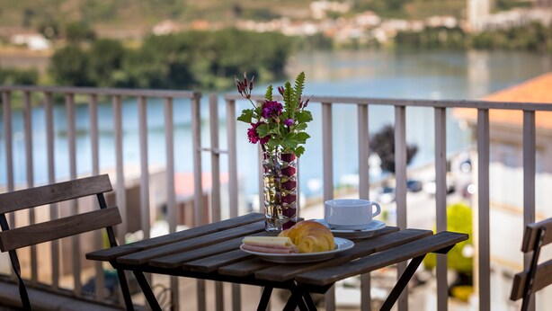 A wooden table with a plate of food, a cup, and a vase of flowers on a balcony overlooking a body of water. - Império Hotel (Peso da Régua)