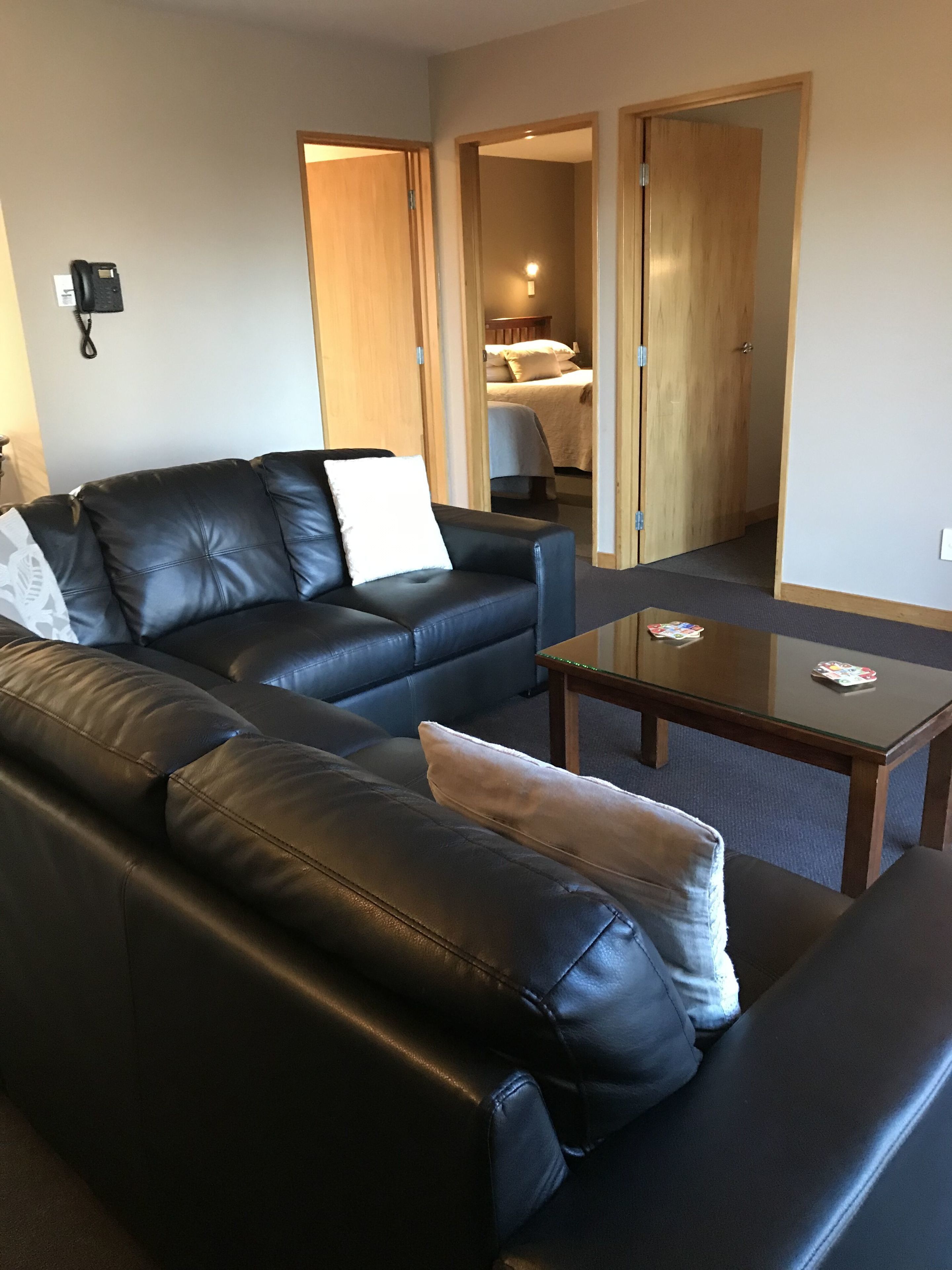 Un salon moderne avec un canapé en cuir noir, une table basse en verre et une porte en bois donnant sur une chambre.