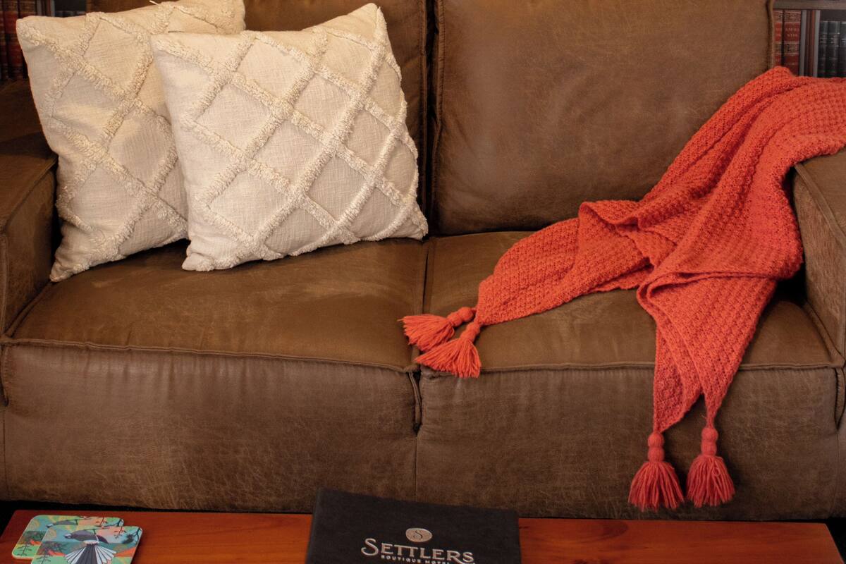 Un sofá de cuero marrón con dos almohadas blancas y una manta roja, una mesa de madera con un libro negro y cartas, y una estantería llena de libros al fondo.