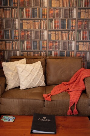 A brown leather sofa with two white pillows and a red blanket, a wooden table with a black book and playing cards, and a bookshelf filled with books in the background.