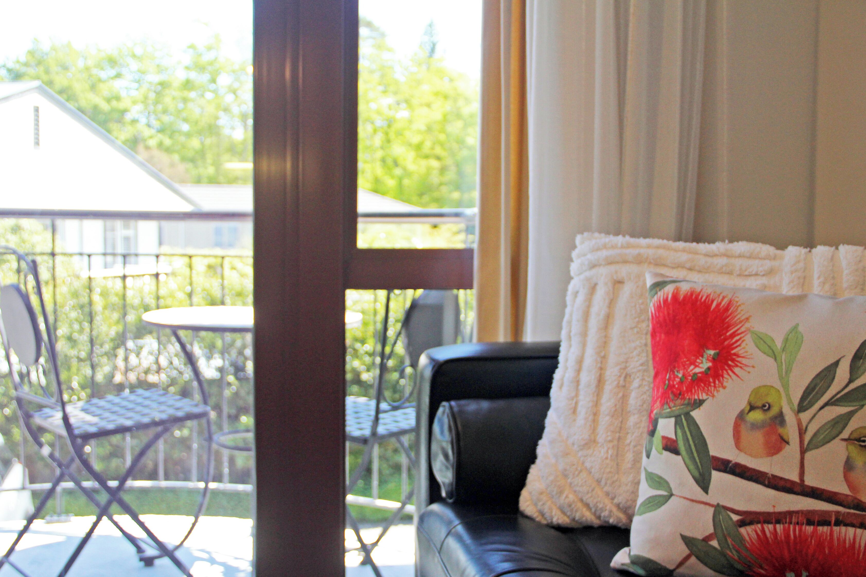 Un balcon donnant sur une maison, un canapé en cuir noir et un coussin orné d’un motif représentant un oiseau et des fleurs.