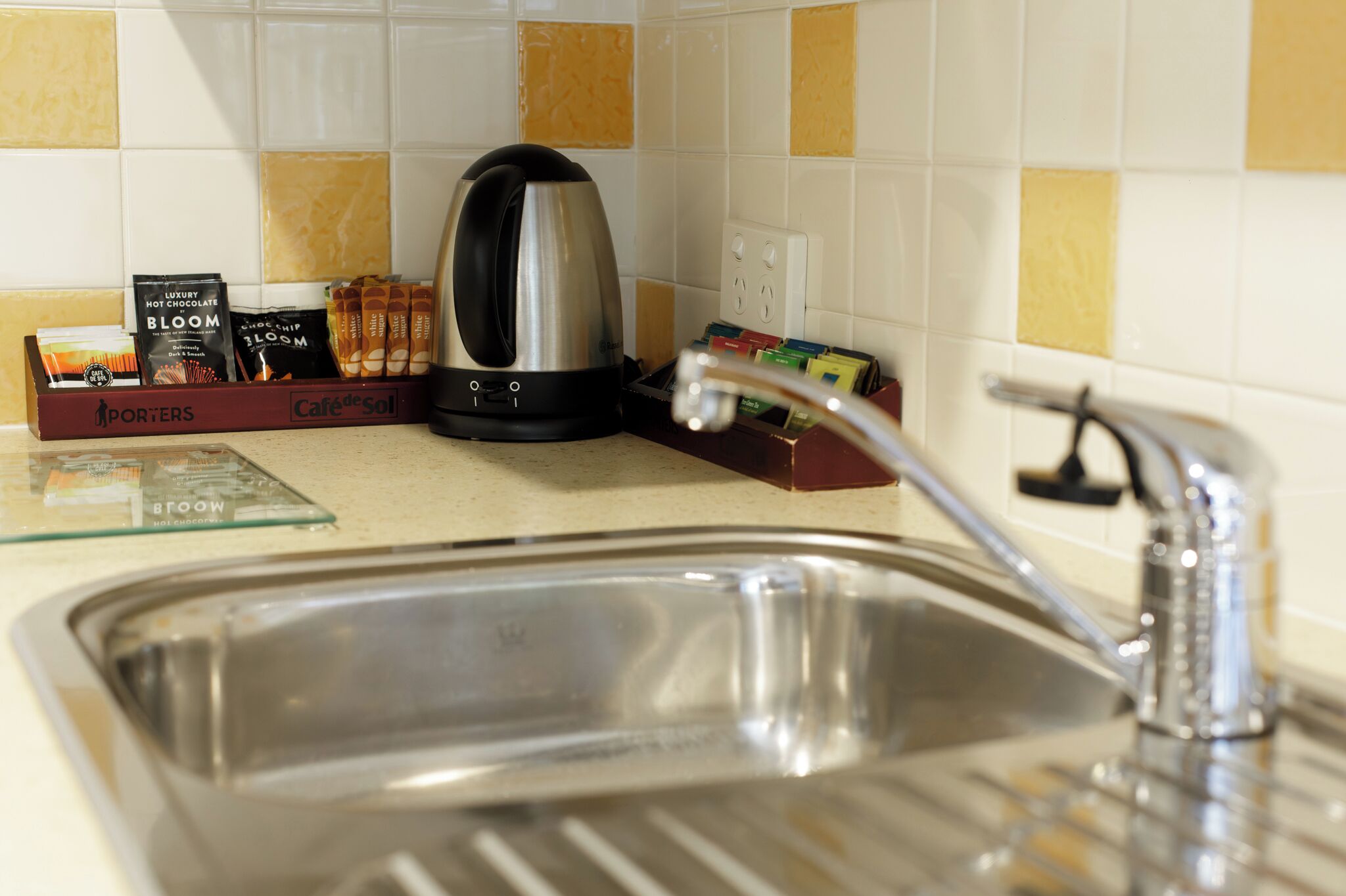 Una cocina con un lavamanos de acero inoxidable, un hervidor y una cafetera. Hay cajas de café y té en la encimera.