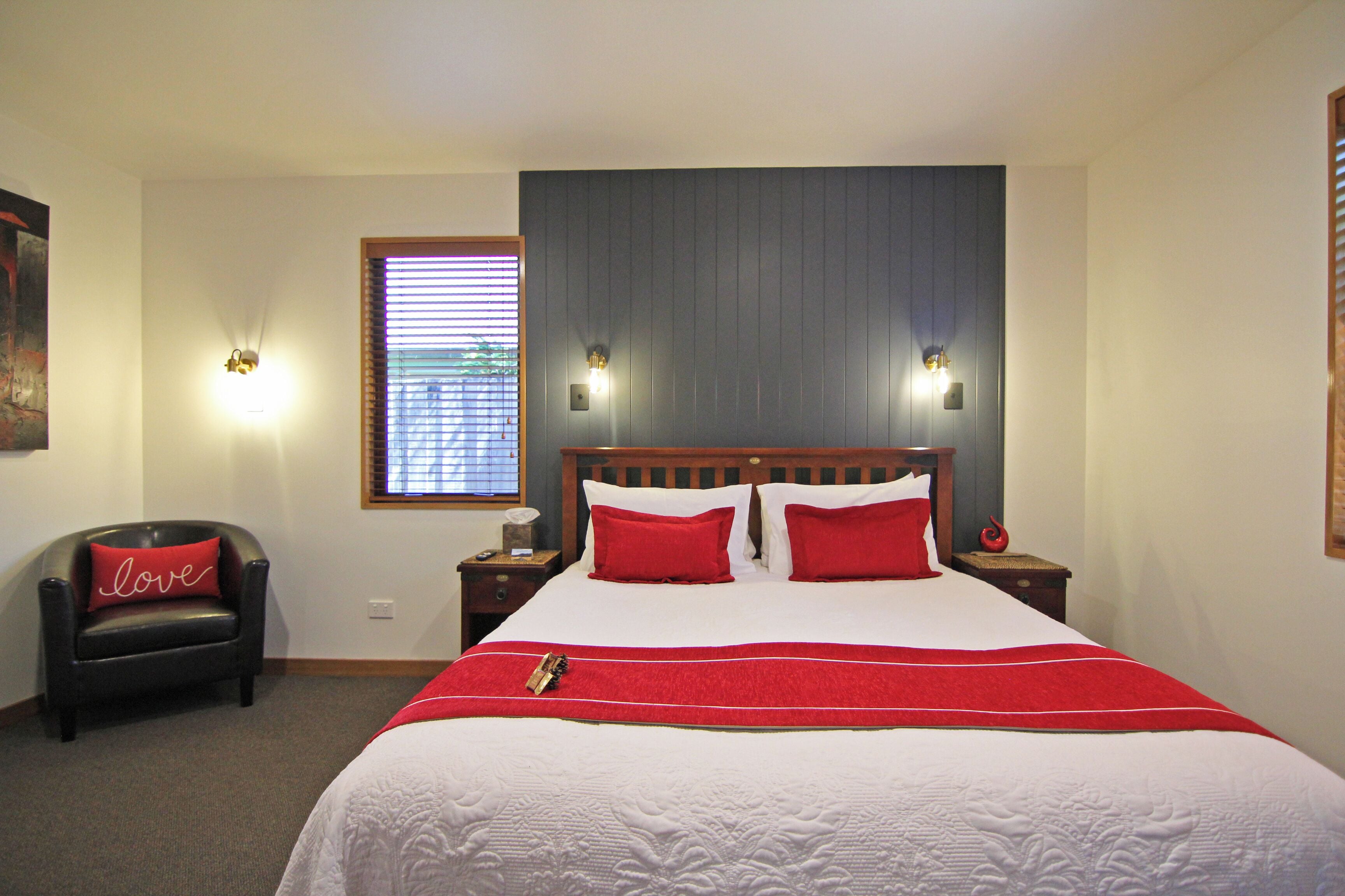 Une chambre d’hôtel avec un grand lit, deux tables de chevet, une chaise, une fenêtre avec des stores et des lampes fixées au mur.