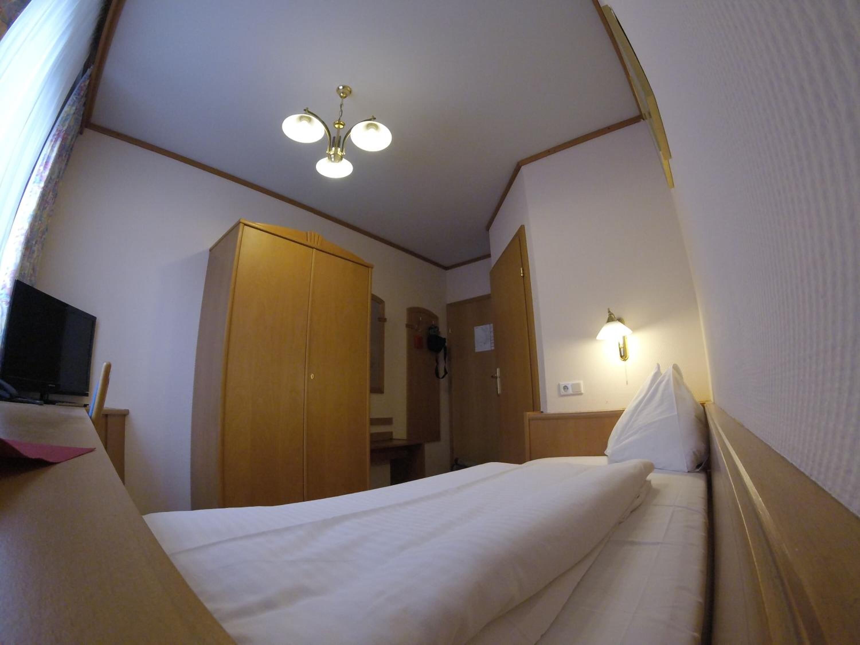 A compact hotel room with a single bed, a wooden wardrobe, a television, and a ceiling light fixture.