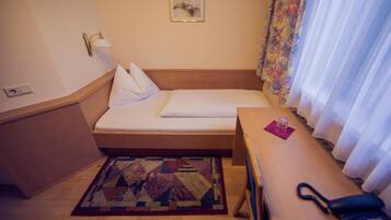 A small, neatly arranged room with a bed, a desk, a telephone, and a patterned rug.