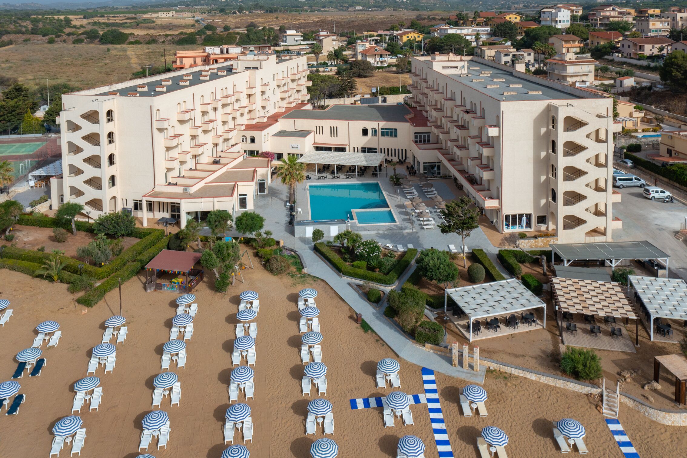 A resort with a central pool, multiple balconies, and a beach area with sun loungers and umbrellas.