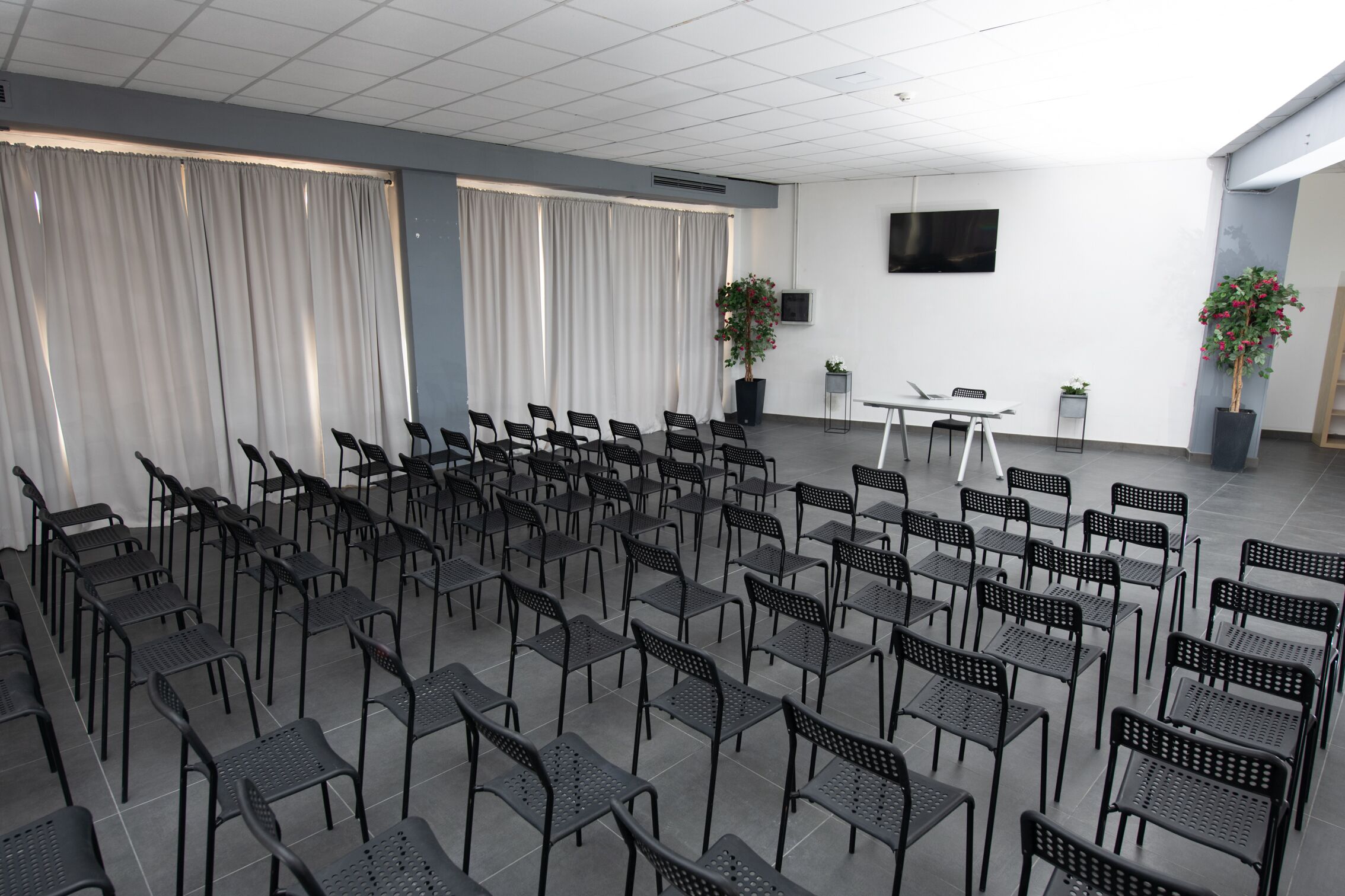 Une salle de conférence avec des rangées de chaises, une table, des plantes en pot et un téléviseur à écran plat accroché au mur.