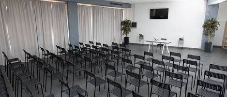 Une salle de conférence avec des rangées de chaises, une table, des plantes en pot et un téléviseur à écran plat accroché au mur.