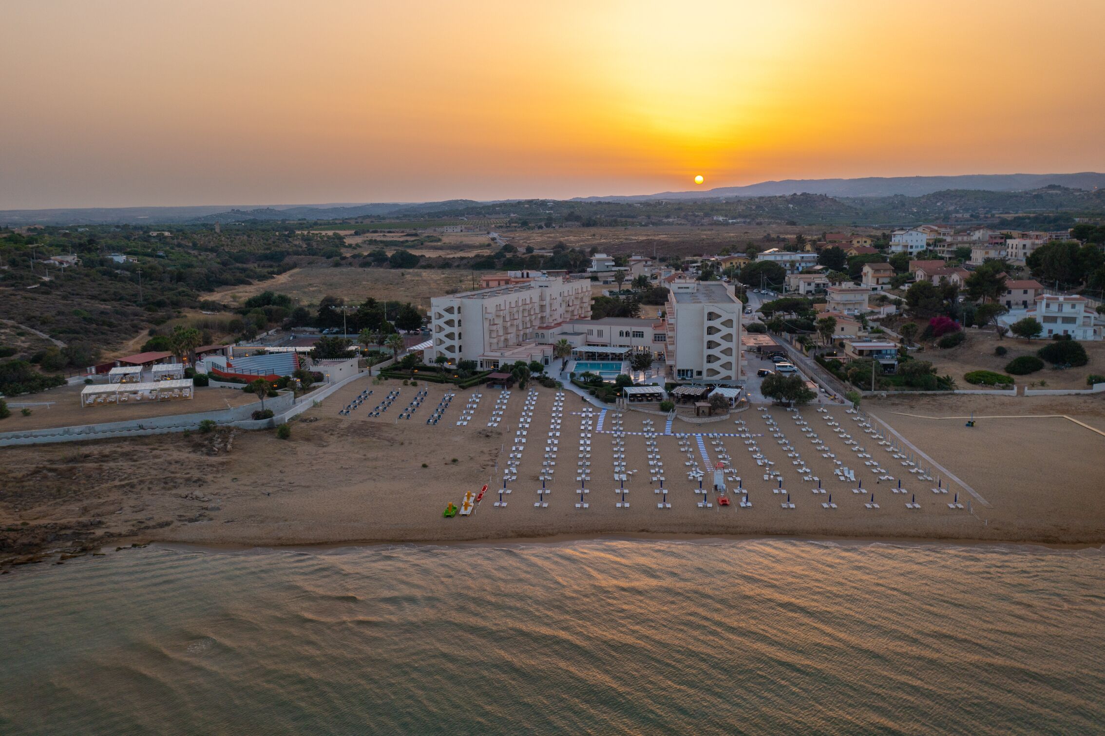A coastal hotel with a large beachfront patio, multiple buildings, and a sunset view.