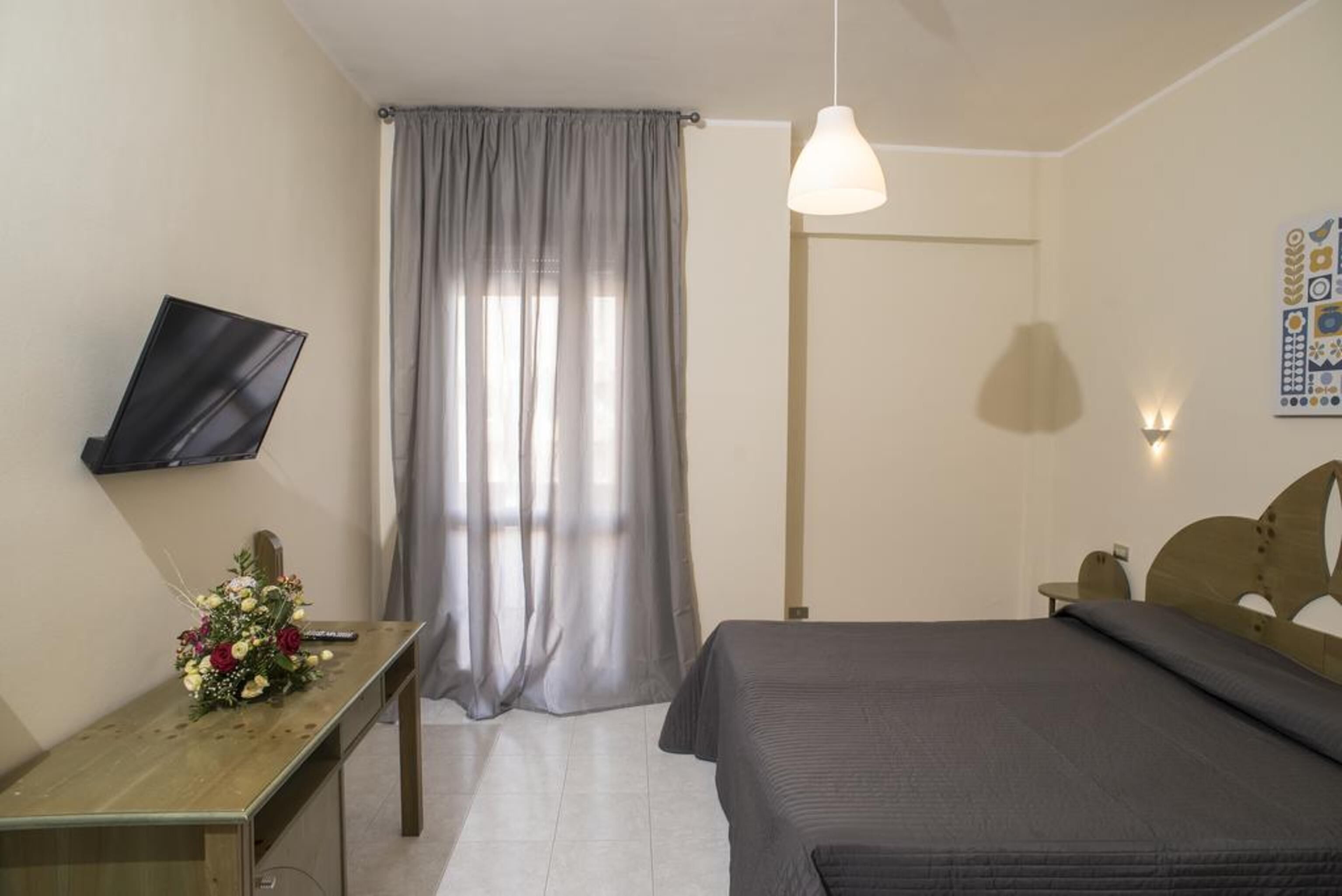 A bedroom with a bed, a desk with a flower arrangement, a TV mounted on the wall, and a window with curtains.