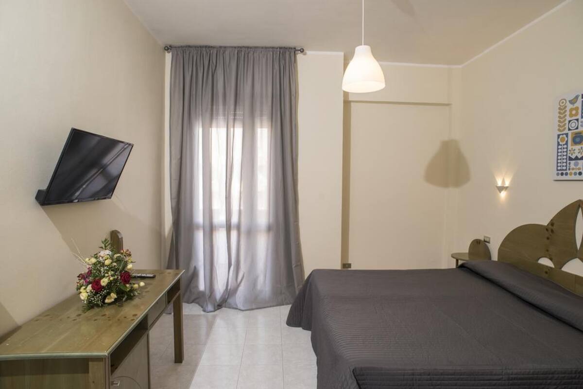 Een slaapkamer met een bed, een bureau met bloemen, een aan de muur bevestigde televisie en een raam met gordijnen.