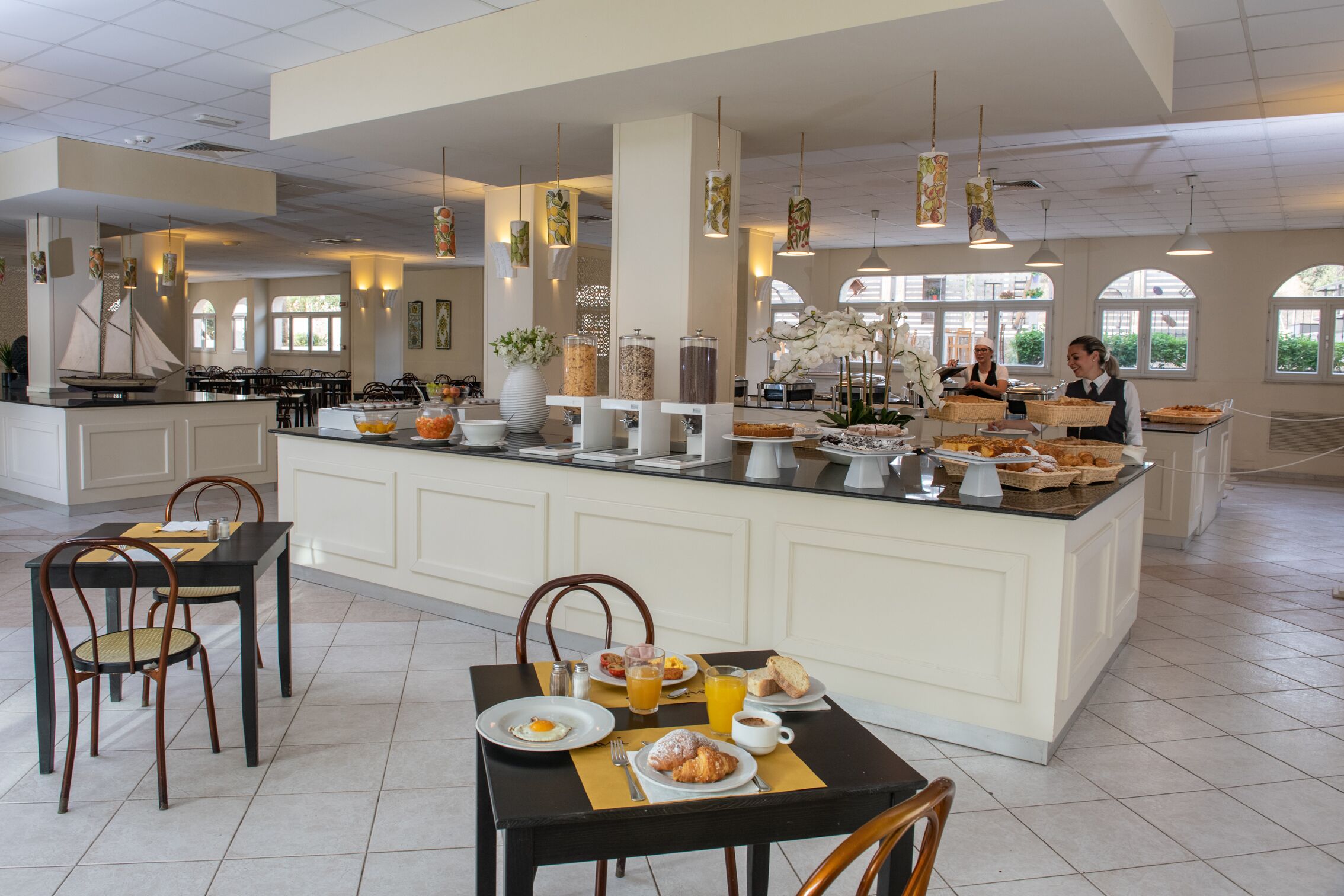Un espace buffet pour le petit-déjeuner avec un îlot central, des tables, des chaises et une vue sur l’extérieur grâce à de grandes fenêtres.